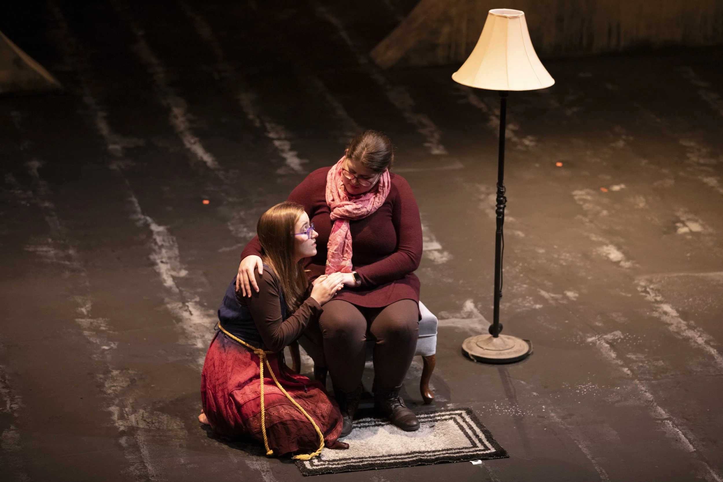 Two women are seated on a stage, engaged in a theatrical performance. One woman, with glasses, has her arm around or touching the other woman, who is sitting on a chair and appears emotional. A floor lamp is positioned nearby. The background is dark 