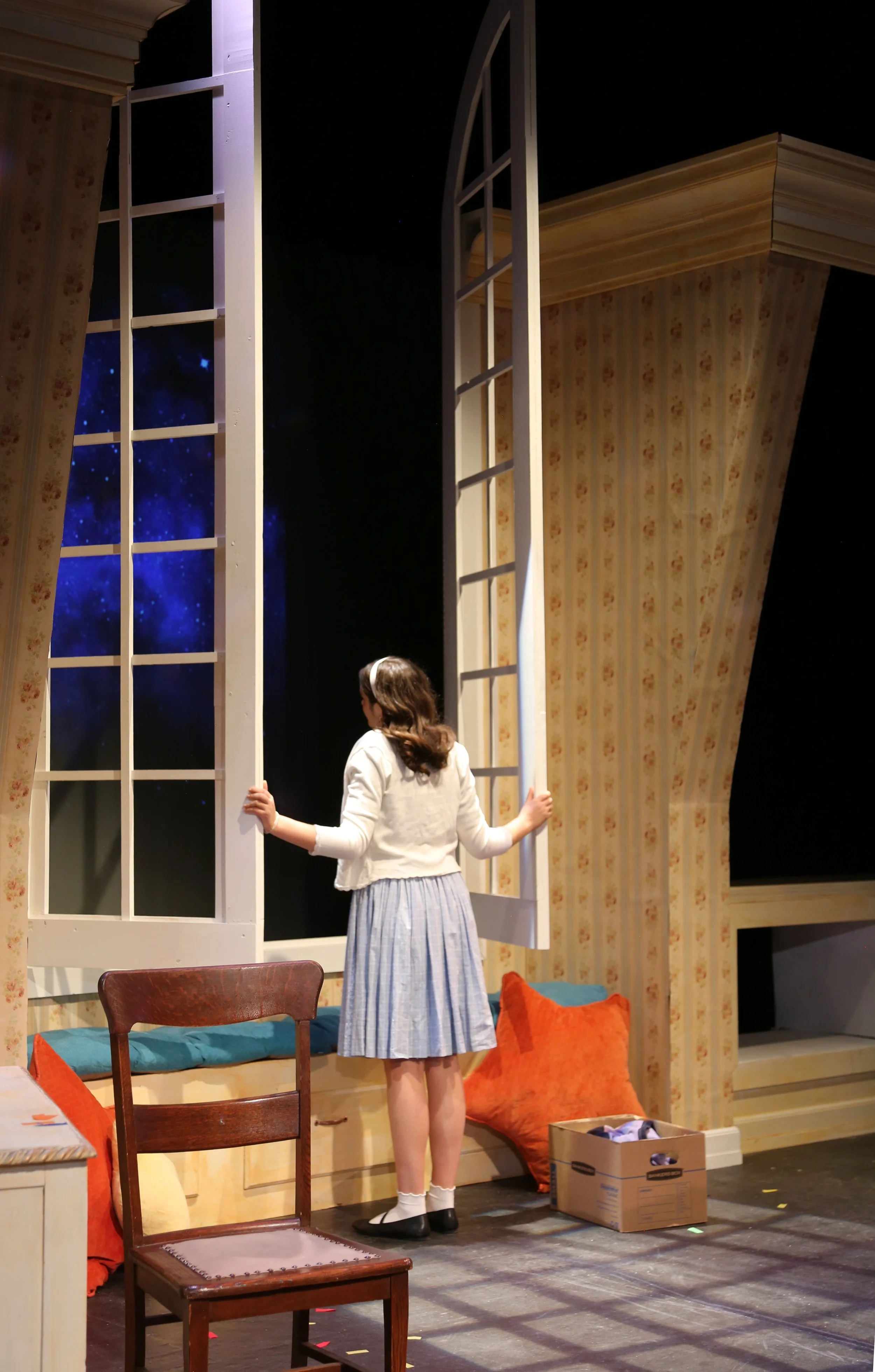 A girl with brown hair in a ponytail wearing a white cardigan and a long blue striped skirt standing inside a room near an open window, looking outside. The room has floral wallpaper, a wooden chair with a beige cushion and orange pillow, and a cardb