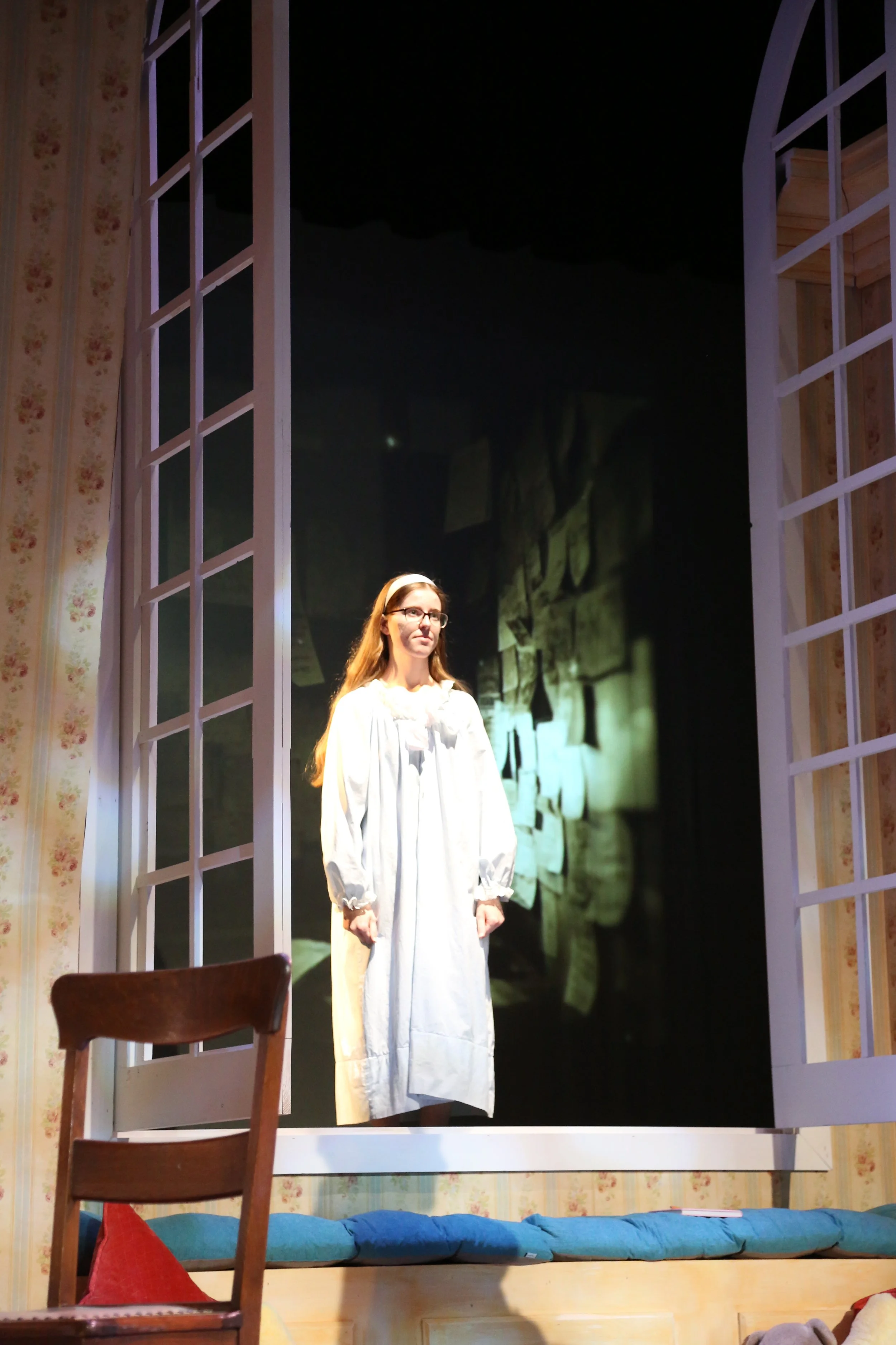 A woman dressed in a long white nightgown stands on a stage framed by an open door with white lattice and floral wallpaper. The scene is part of a theatrical performance or play.