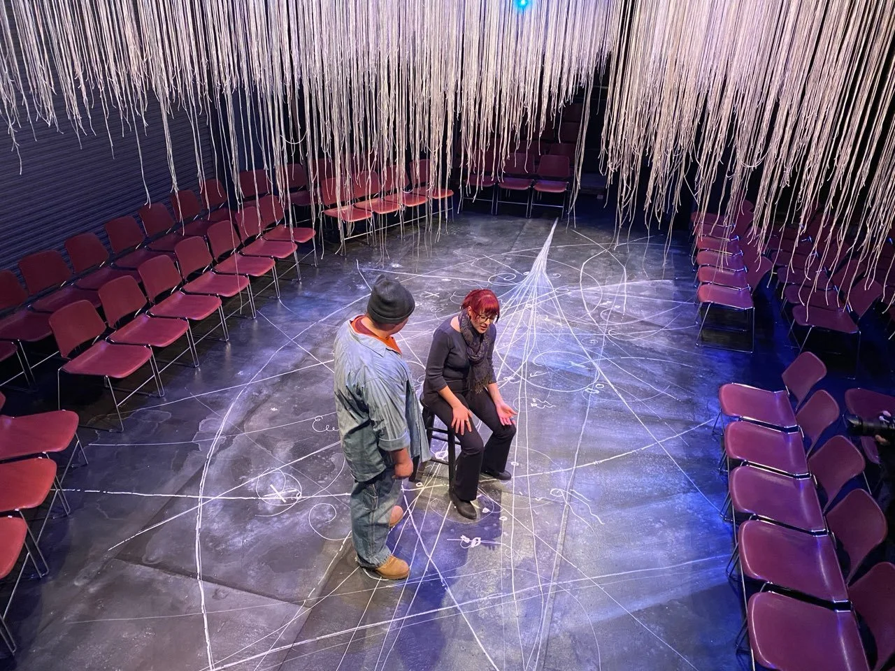 Two people are in an intimate stage setting with hanging strings and chalk outlines on the floor, surrounded by rows of purple chairs.