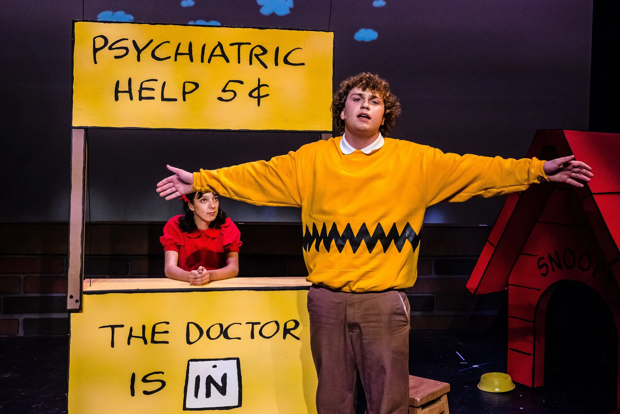 A theatre scene from 'You're a Good Man Charlie Brown', with a young man dressed as Charlie Brown standing in front with arms spread wide, Lucy sitting behind a yellow desk with a sign that reads 'Psychiatric Help 5¢'.