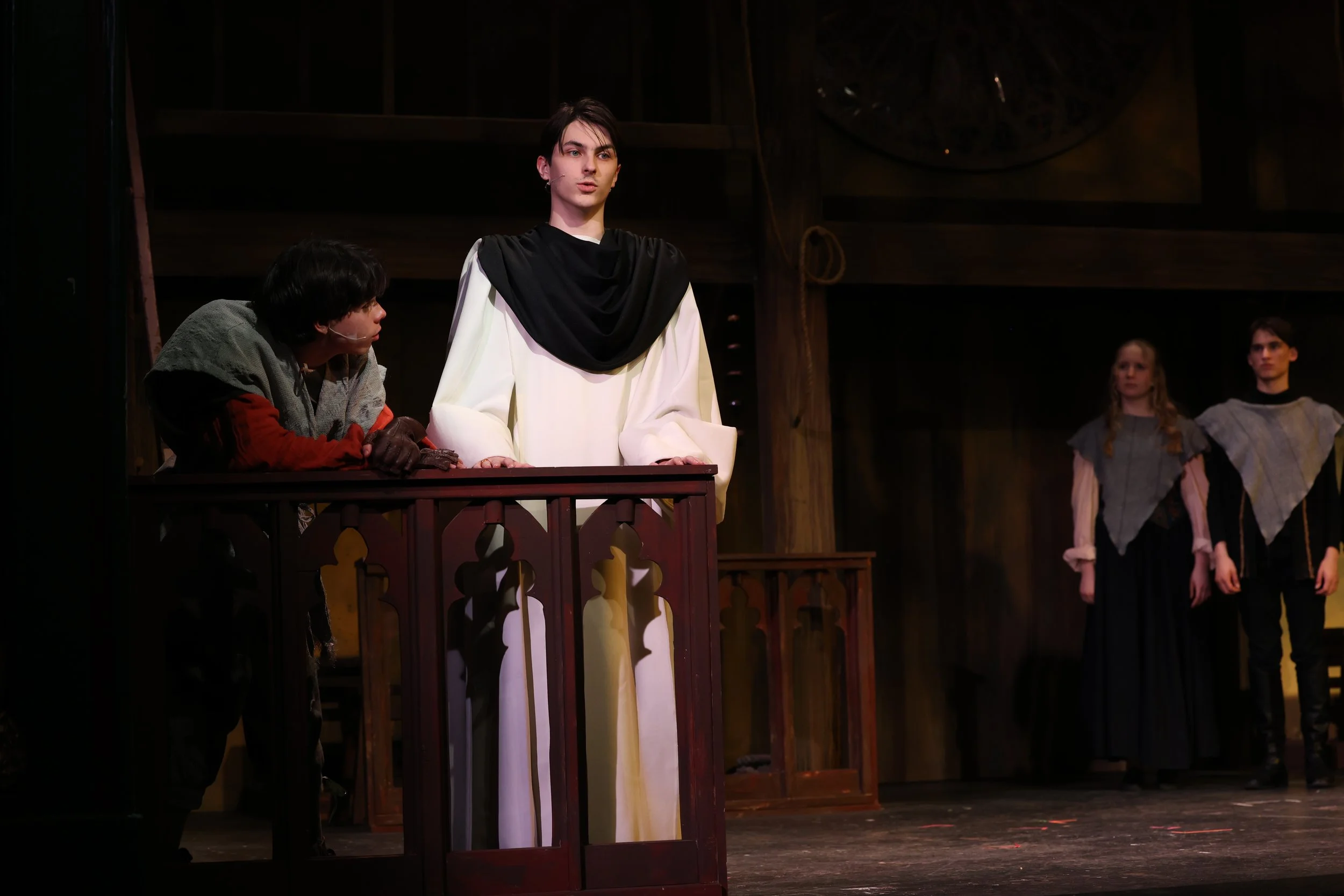 Actors performing on stage in a theatrical play, with one actor in white and black robes at the center and others in shadow or costumes to the side.