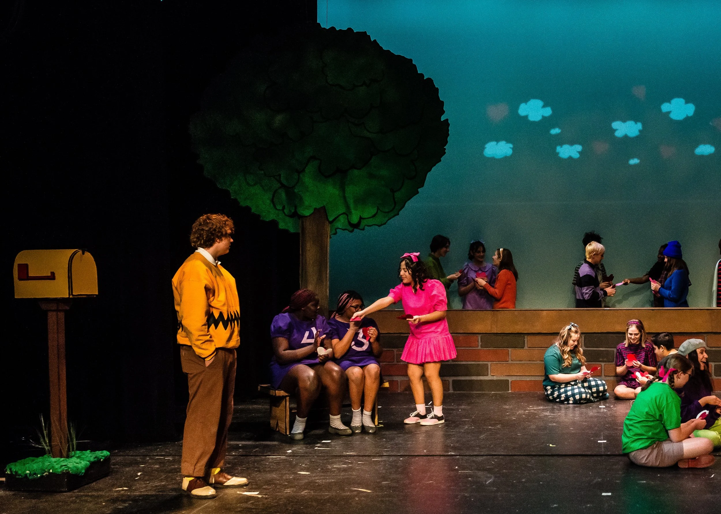 People on stage, some seated and some standing, with a large paper tree and clouds in the background. An actor playing Charlie Brown is on the left, and others are dressed in colorful costumes, engaging in a performance.