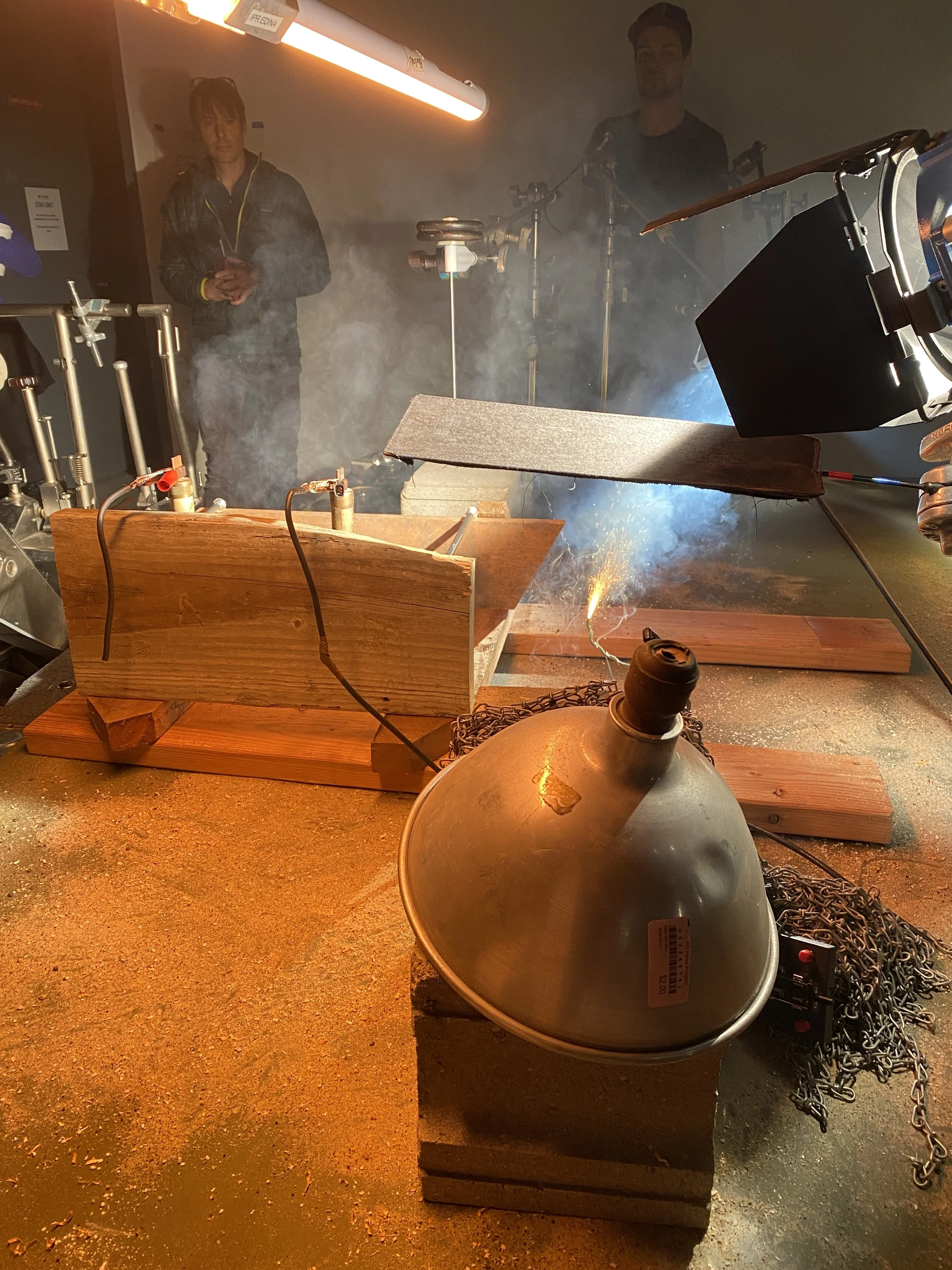 A film crew with a Rube Goldberg-style setup, with a wooden box and metal equipment. Sparks are visible, and two people are in the background observing. Both are standing in a smoky environment with various machinery around.