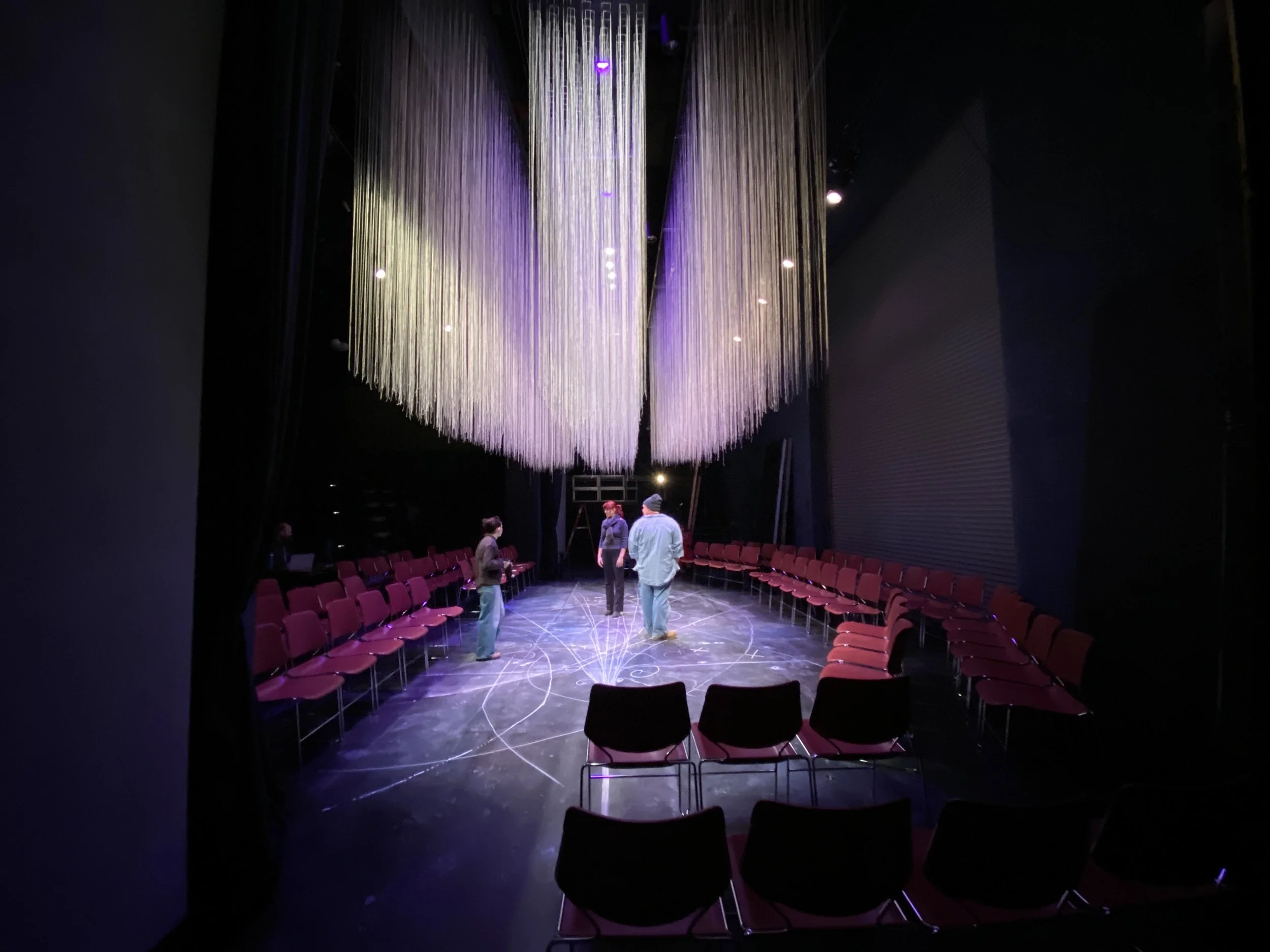 Interior of a theater with a large installation of strings hanging from the ceiling, rows of red chairs, and a group of people standing in the center.