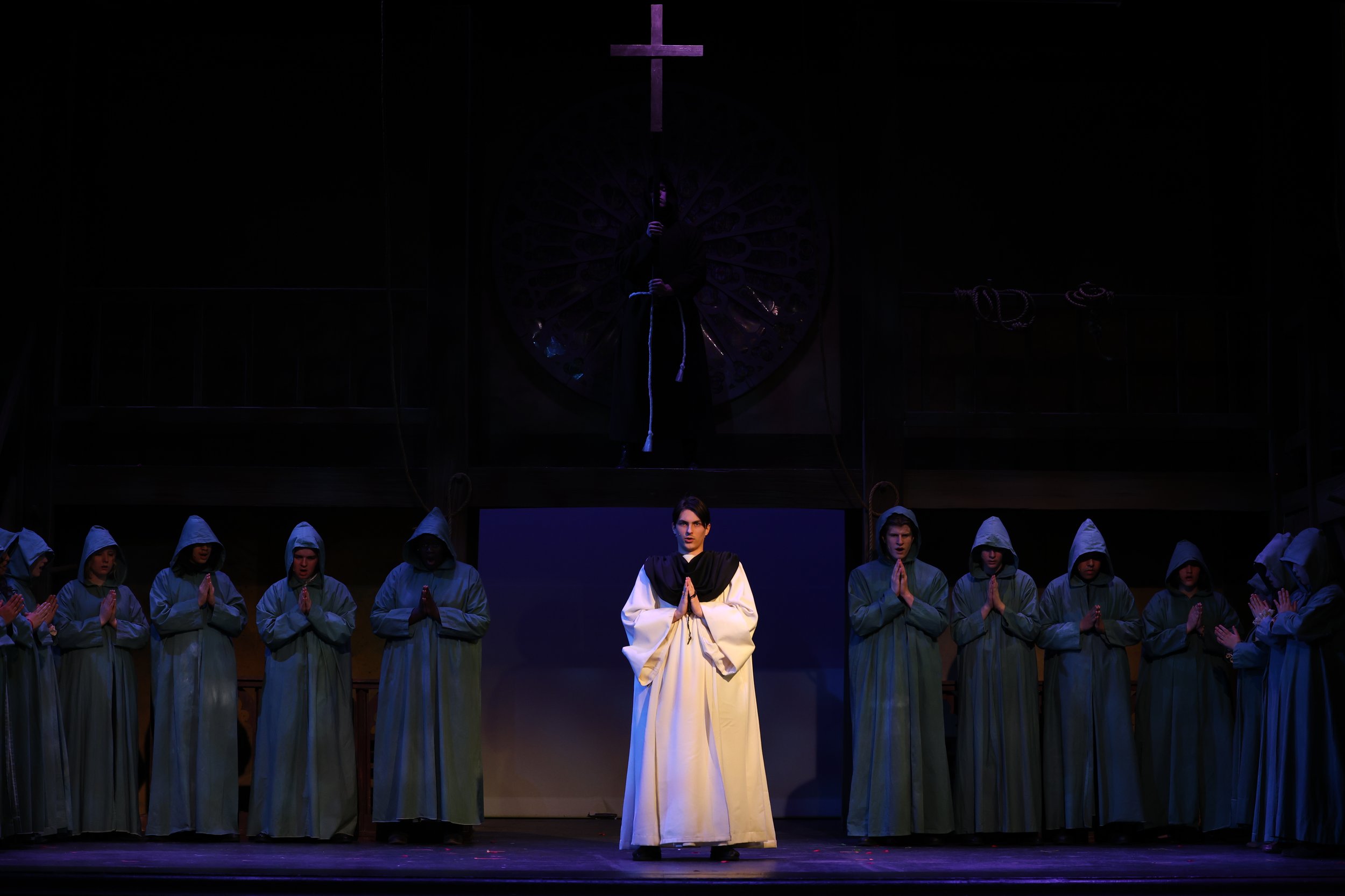 A theatrical performance with a group of people dressed in robes on stage, with one person in front wearing a white robe and others in green robes. Behind them, a person is holding a cross above their head, with the scene dimly lit in a dark setting.