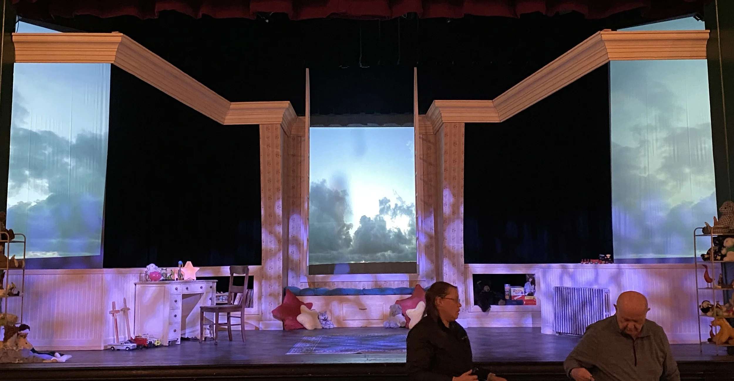 A stage decorated with a large backdrop of a cloudy sky and a wooden frame resembling a house. There are children's toys and furniture on the stage, including a small white desk, chairs, and stuffed animals. Several people are present near the stage,