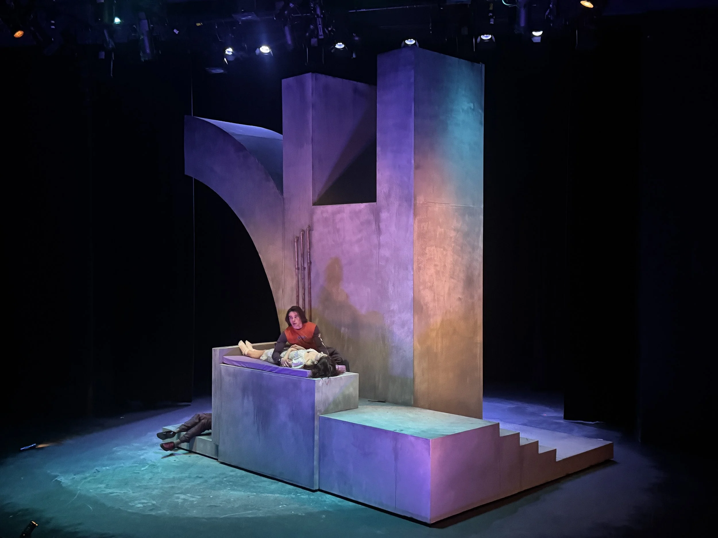 A theatrical stage scene with pastel-colored geometric set pieces including a large block, a staircase, and platforms. A woman is lying on the stage floor near the set, looking up, and another person is sitting on a platform, partially visible at the