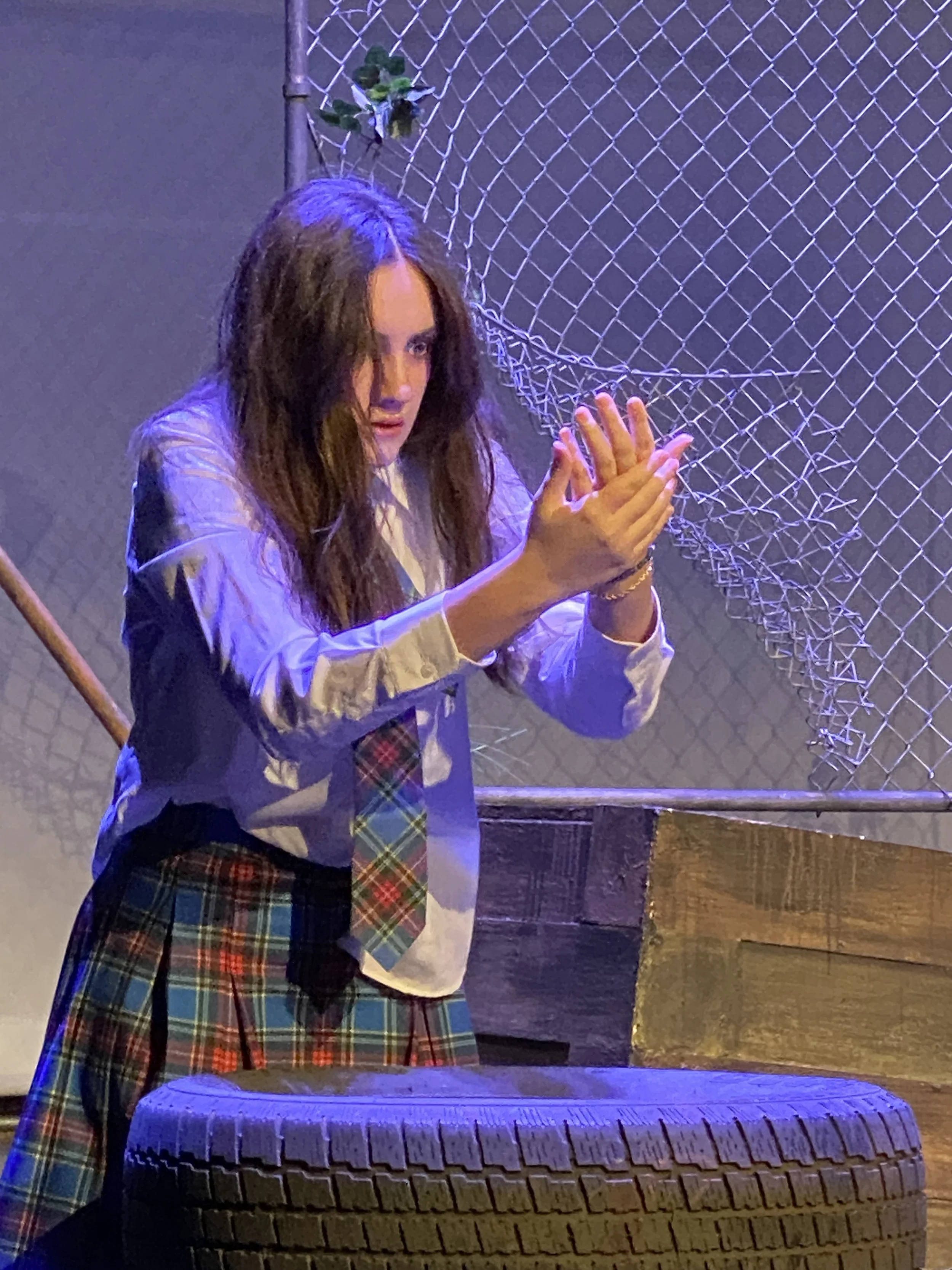 A woman with long brown hair wearing a school uniform with a white shirt, plaid tie, and plaid skirt, standing near a chain-link fence and a stack of tires, with purple and blue lighting.