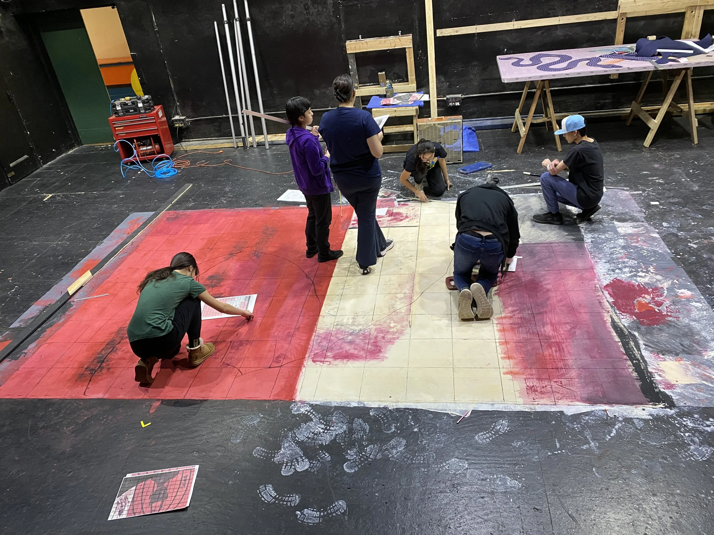 People working on a stage set, painting and preparing a large red and beige backdrop, with various tools and materials around them in a black-walled room.