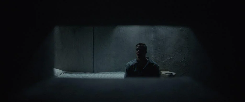 A man sits in a dimly lit prison cell or similar confined space, seen through a small rectangular opening.