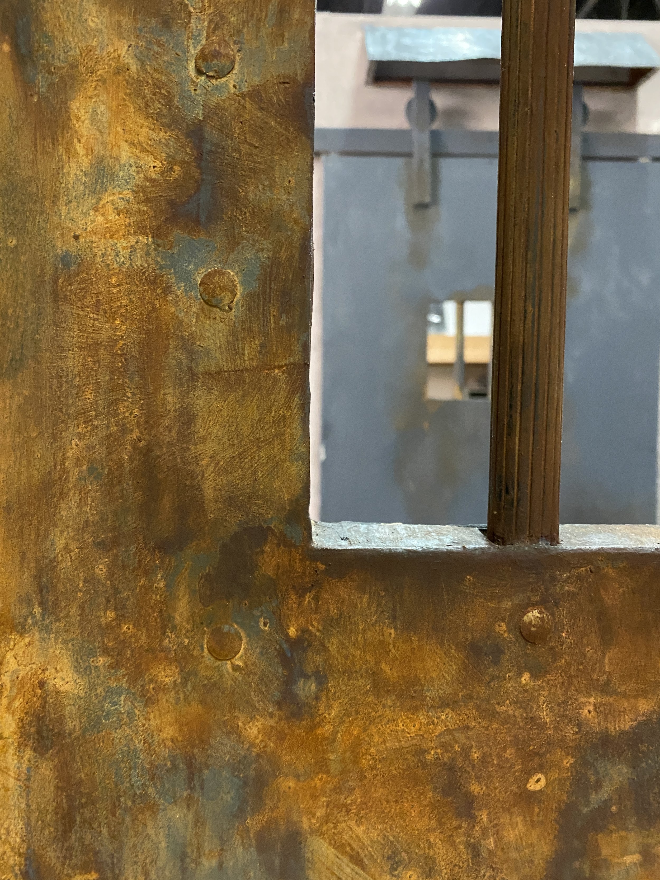 Close-up of rusted metal surface with orange and blue tones, showing the edge of a window or opening with a metal grille or bars behind it.