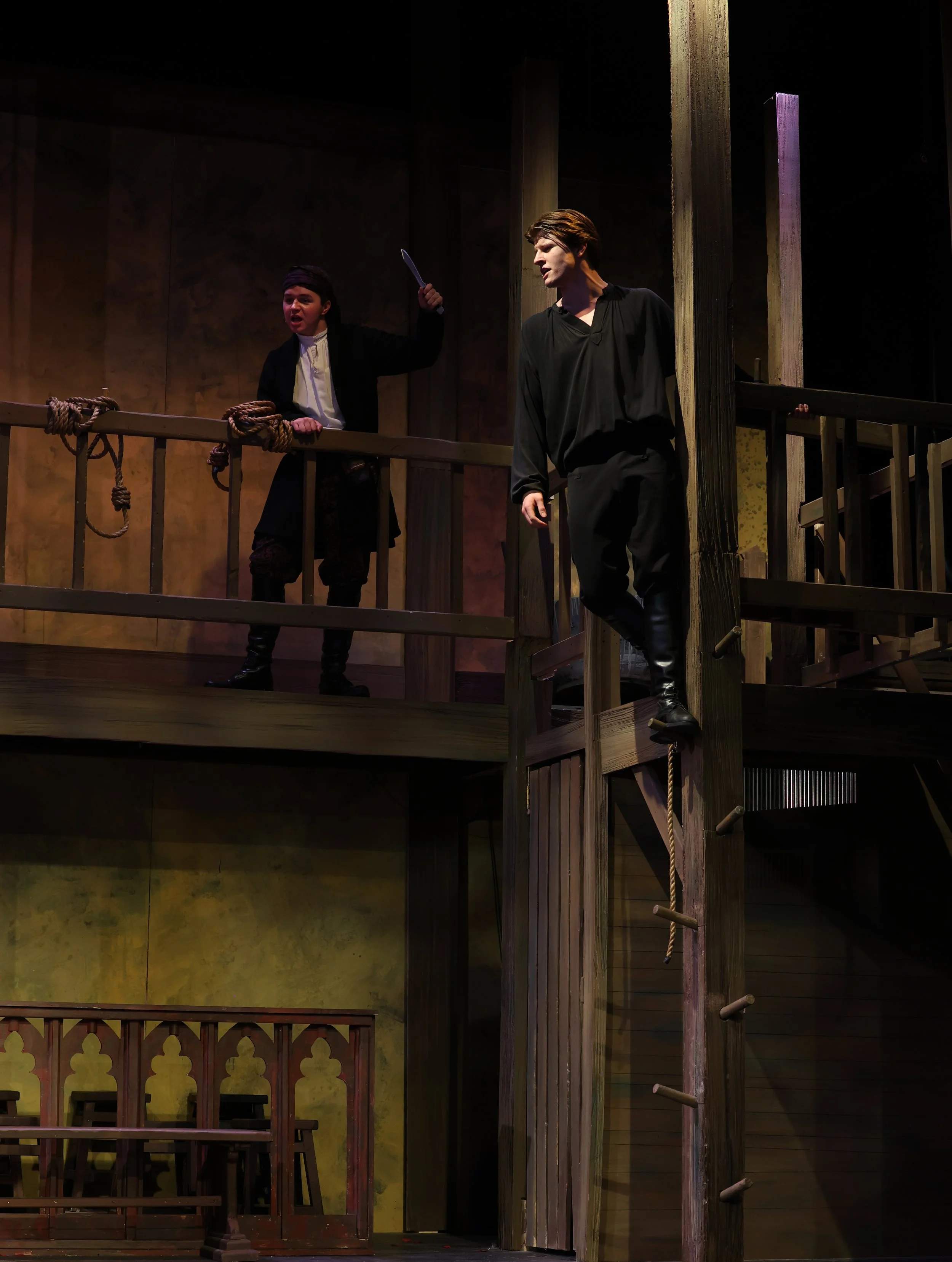 Stage scene from a theatrical play, with two male actors dressed in period costumes. One actor holds a sword and appears to be arguing, while the other stands near a wooden post and looks at him. The set has wooden railings, ropes, and a yellow wall 
