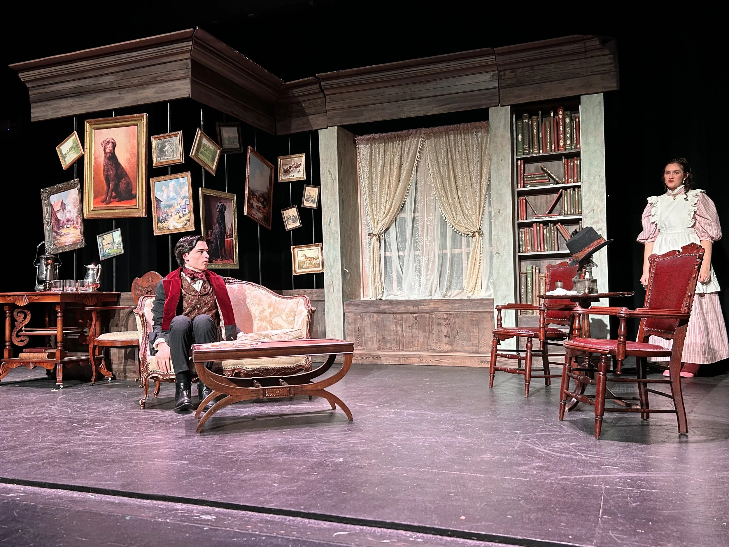 The image shows a stage set resembling a vintage living room, with actors performing. The background has framed paintings, a window with lace curtains, and a bookshelf. The scene includes a woman standing on the right in period costume and a man seat