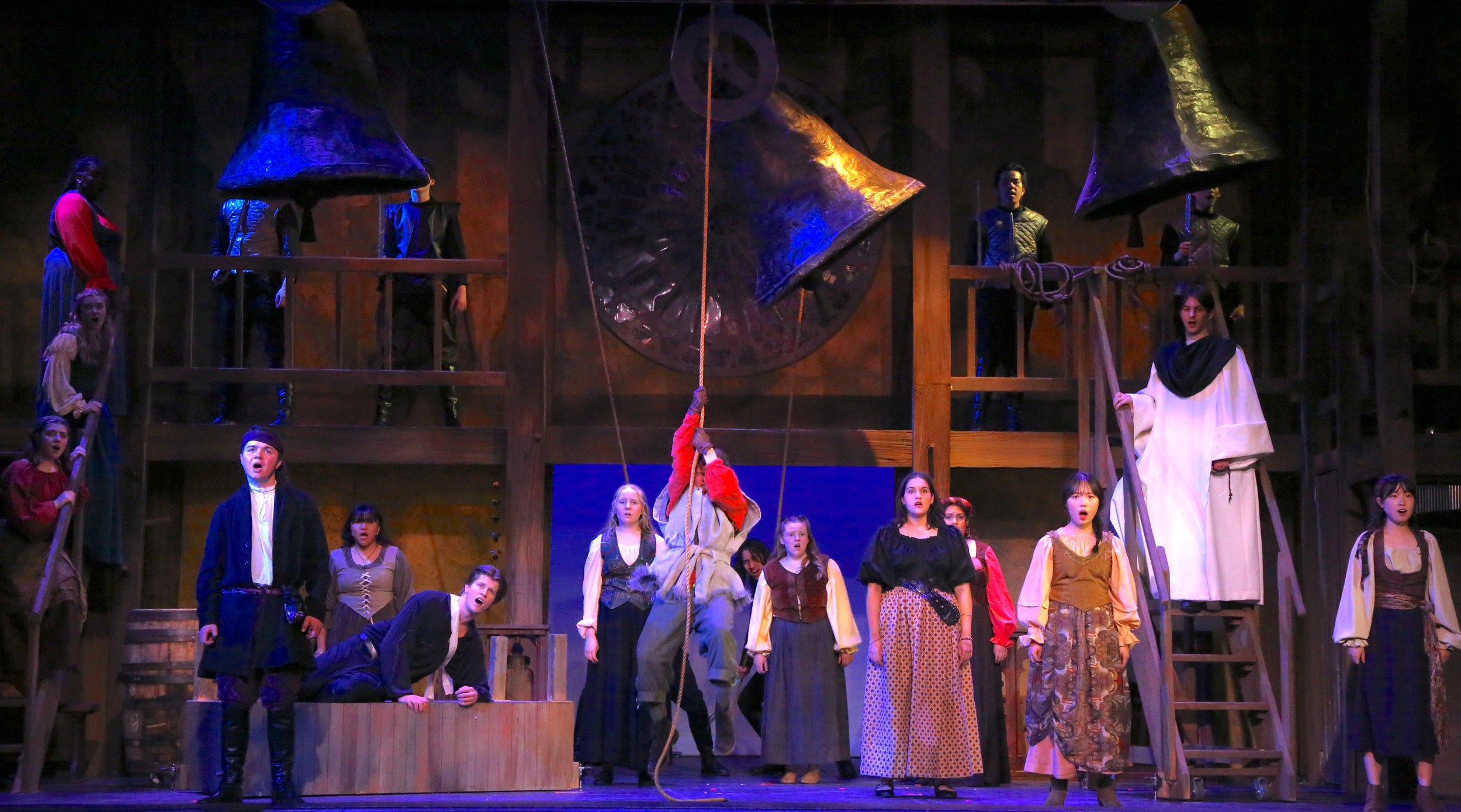 A theatrical stage scene with multiple actors dressed in period costumes, some standing on a wooden structure resembling a cathedral, with a large stained glass window and large bells overhead.