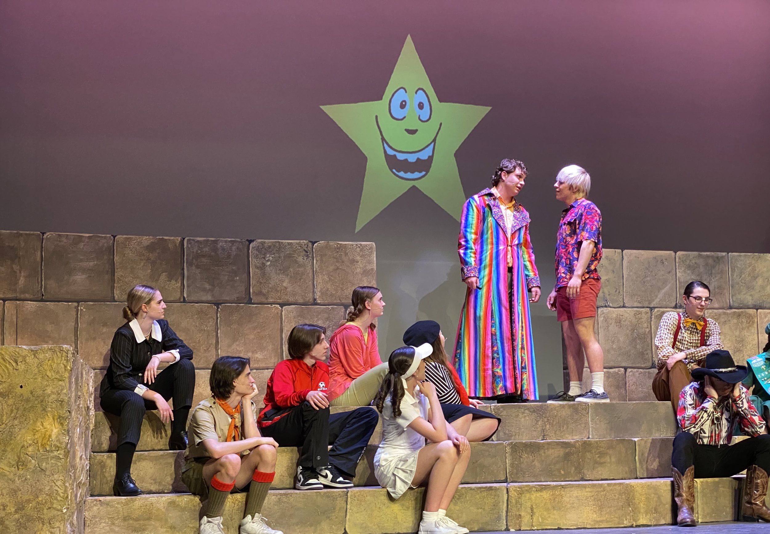 Stage scene with a group of people, some seated on stone steps, others standing, with a large green star with a smiling face projected on the background wall.