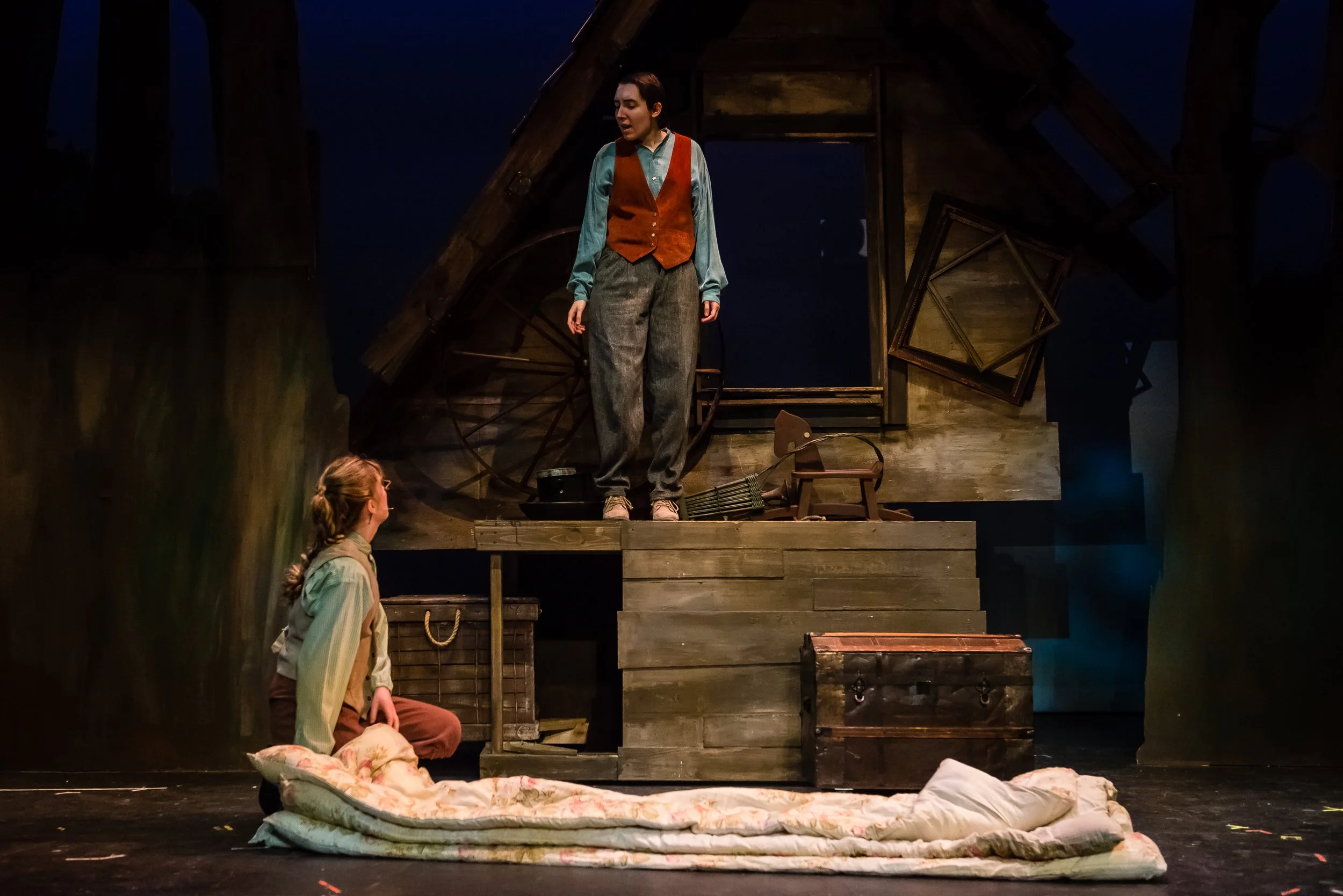 The image depicts a stage scene with three women in period costumes. One woman stands on an elevated platform wearing a brown vest over a turquoise shirt and gray pants, looking contemplative. Two women are seated on the stage; one on the floor near 