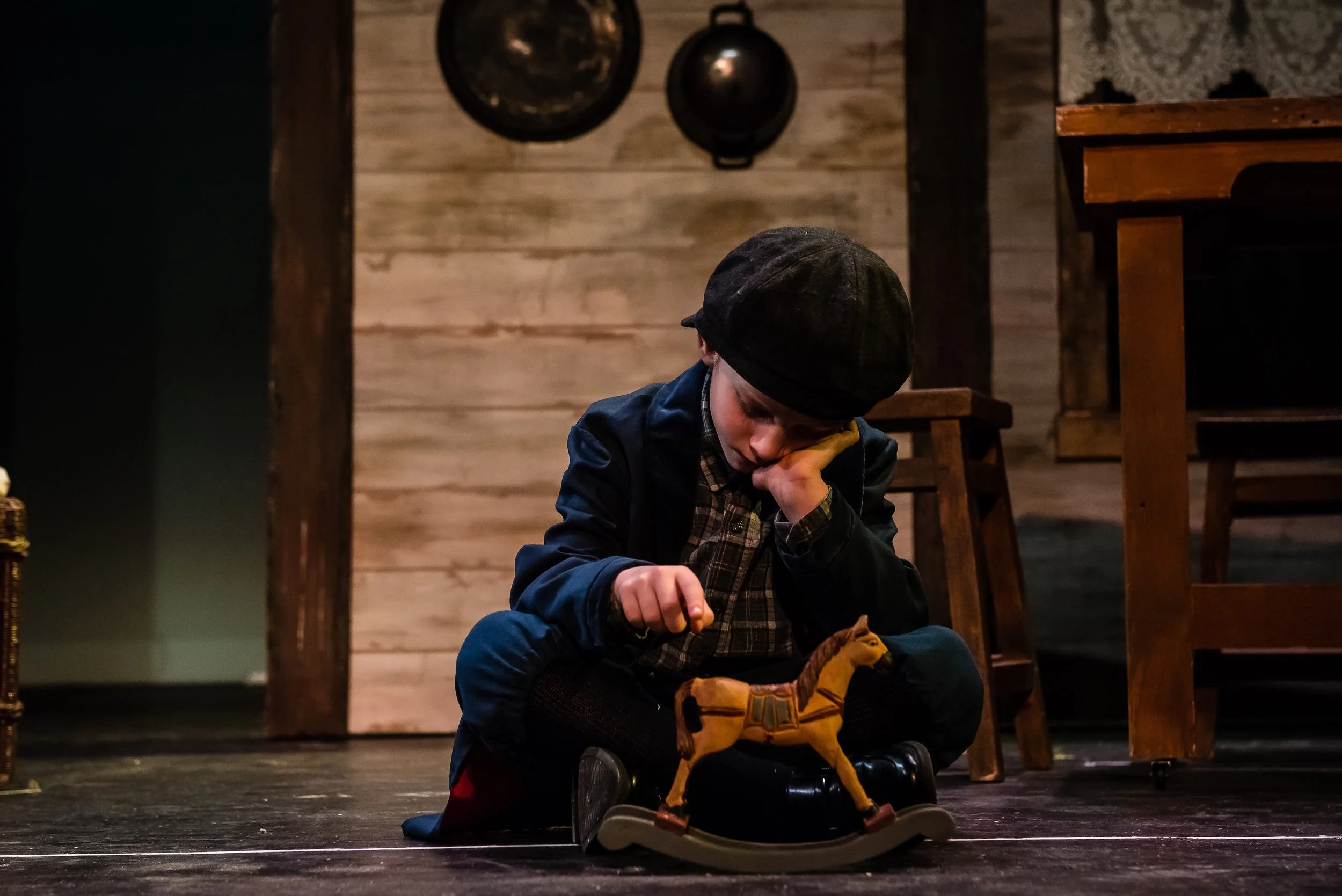 A young boy sitting on the floor of a stage with a wooden background, wearing a flat cap, checkered shirt, dark jacket, and yellow boots, focused on tying his shoelaces.
