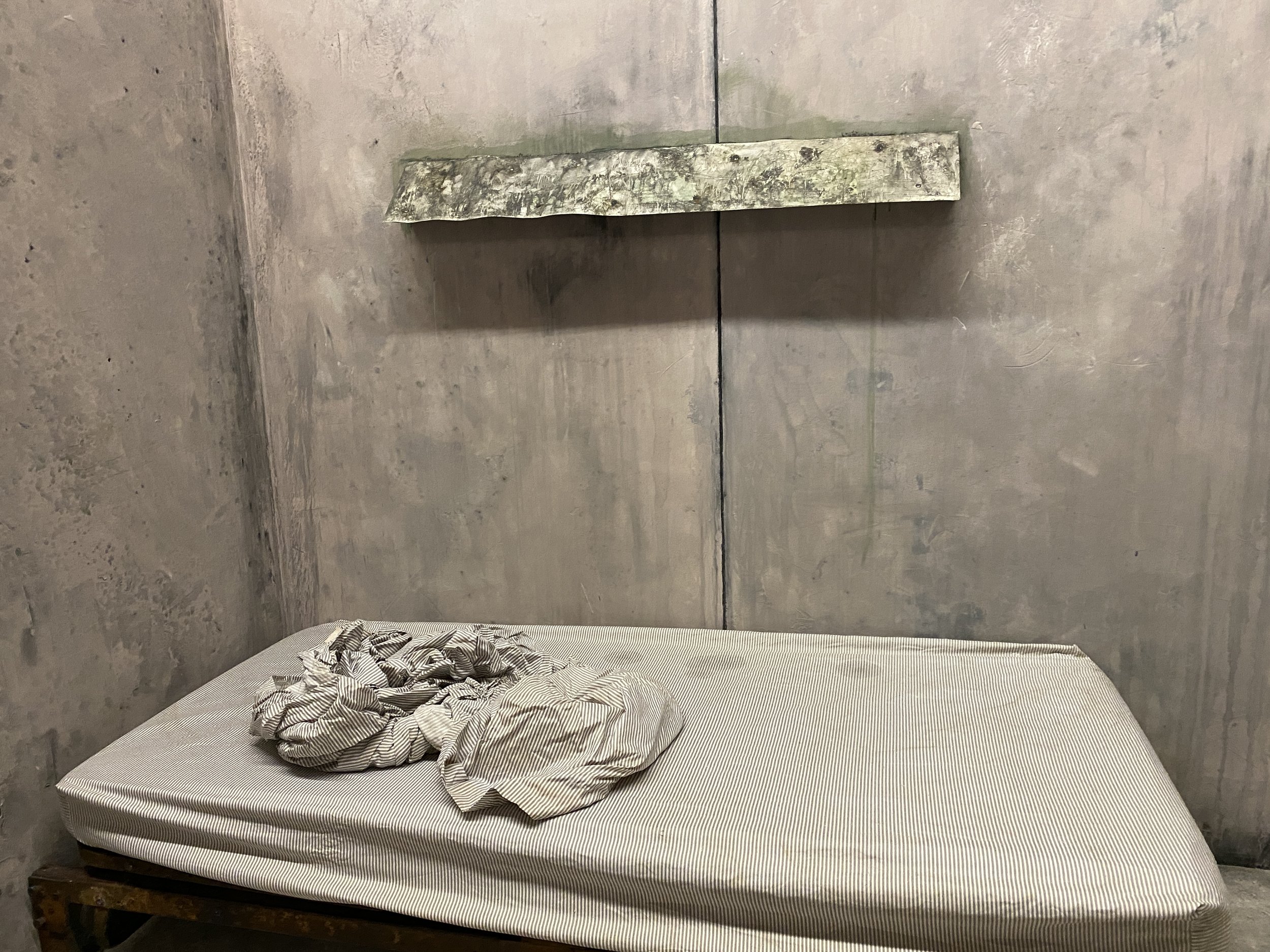 A film set with a bed with a striped mattress and crumpled bedding against a concrete wall in a minimalist, industrial-style room.