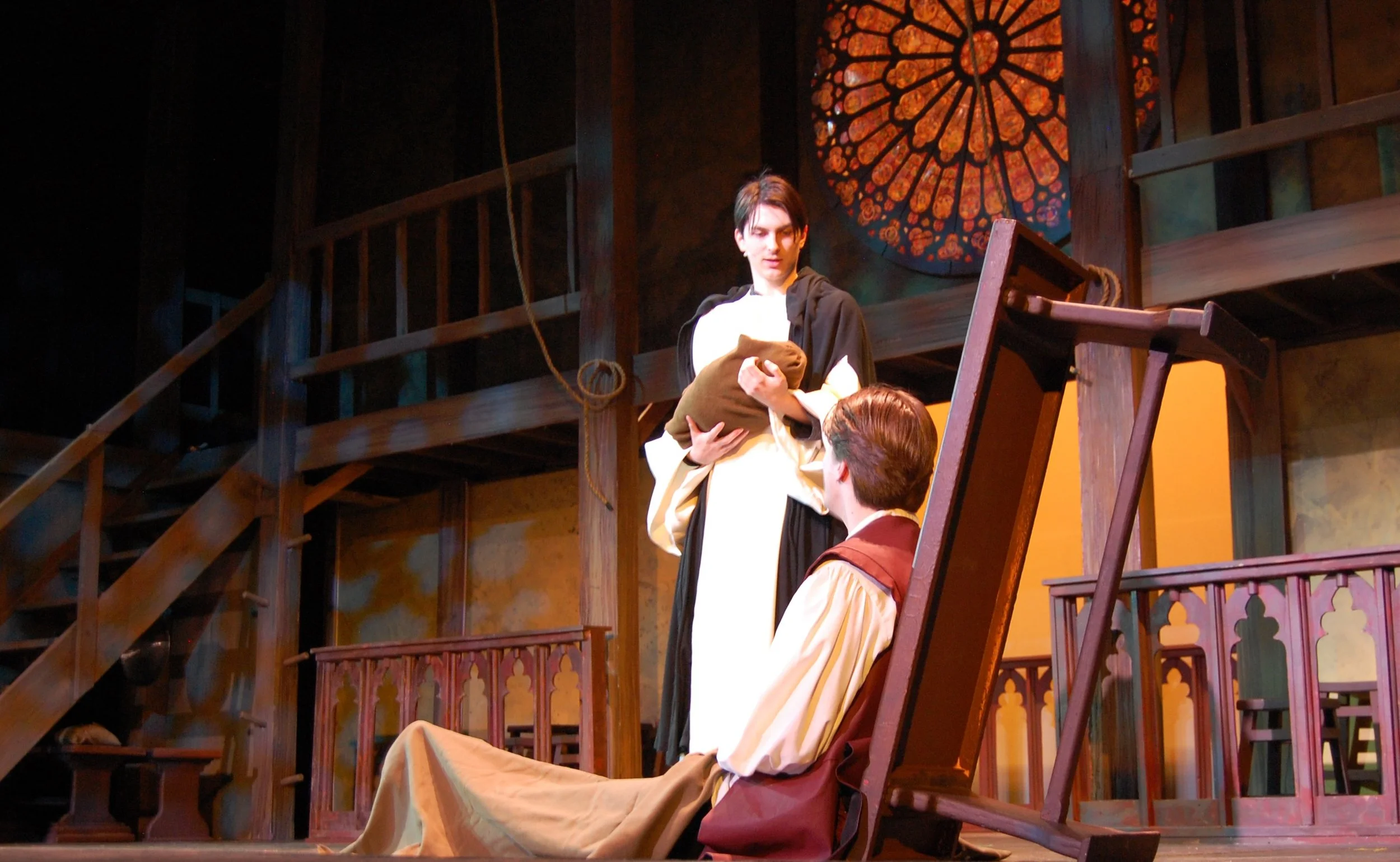Stage scene with actors in period costumes with a large stained glass rose window and wooden staircase in the background.