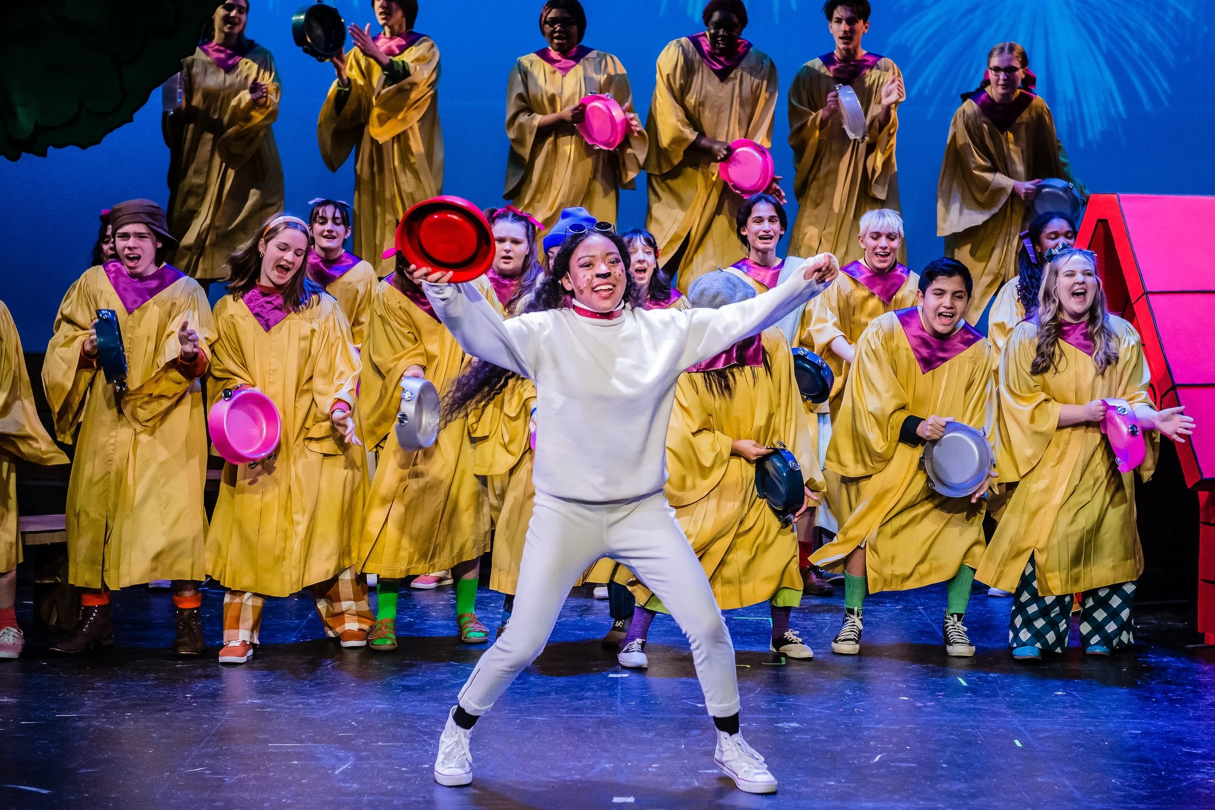 Group of children and a young woman performing on stage in a theatrical production, dressed in colorful costumes with some holding hats and tambourines, smiling and engaging with the audience.