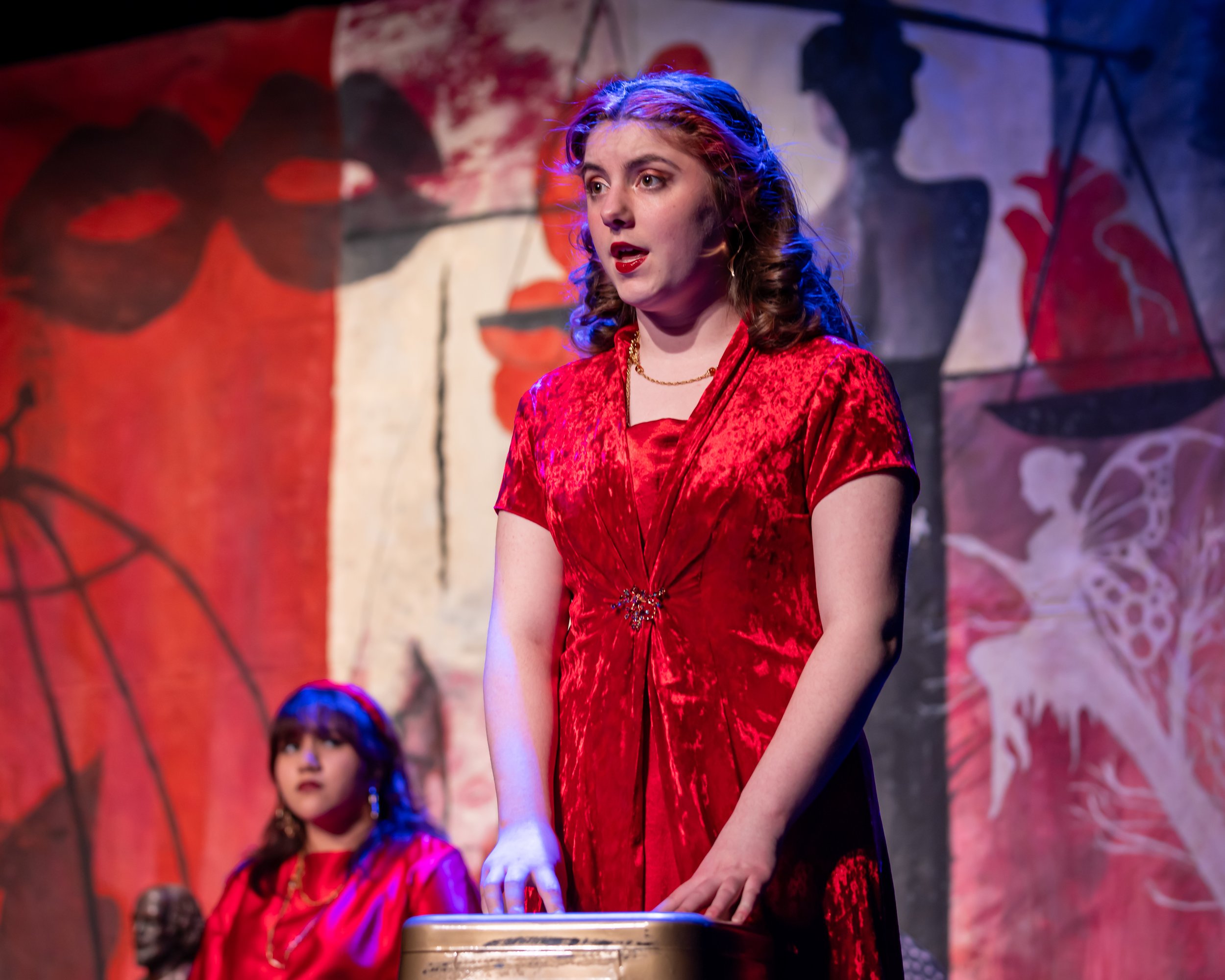 A woman with curly hair wearing a red dress and red lipstick standing on stage during a performance, with a painted backdrop behind her.