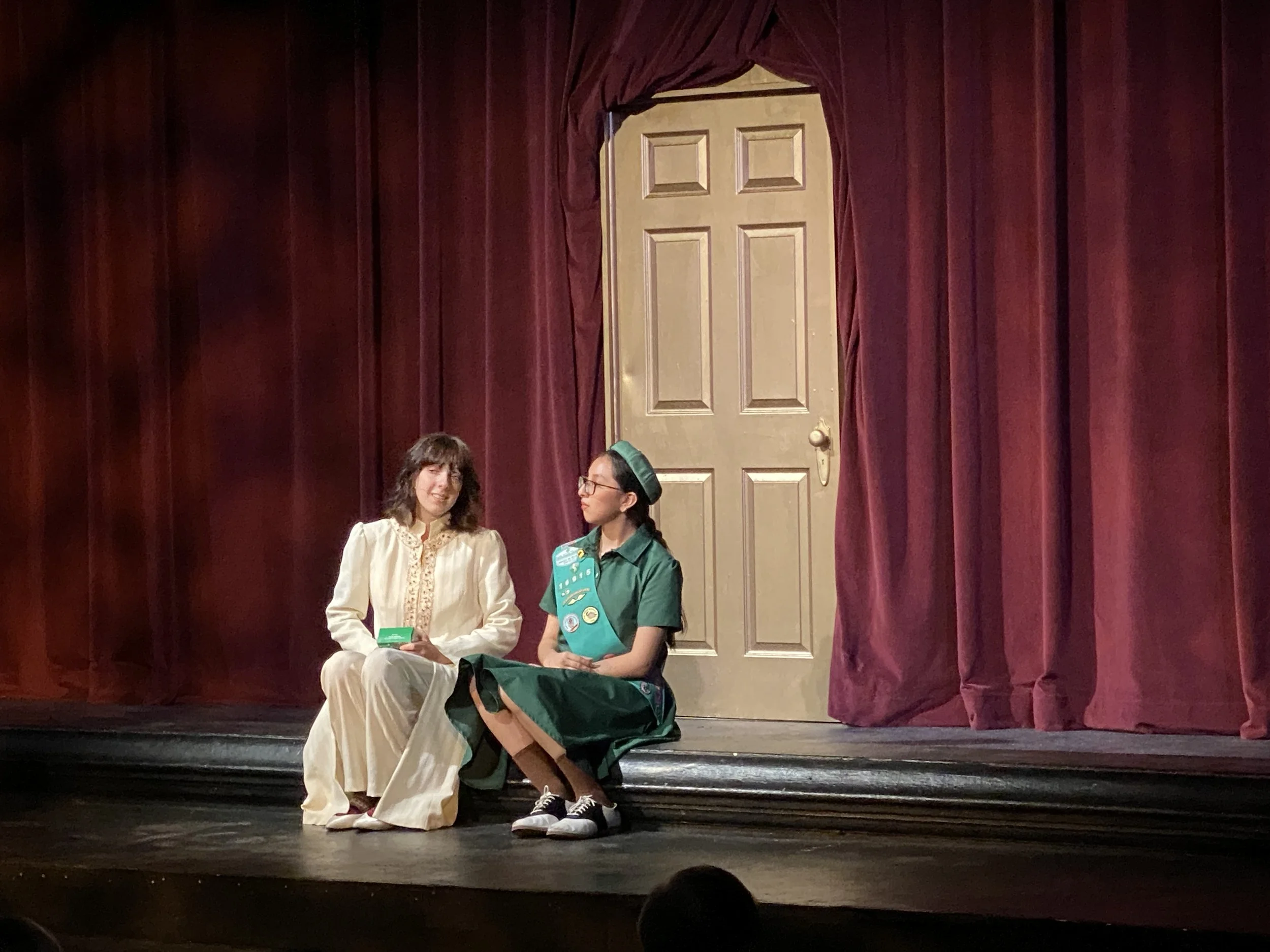 Two women are seated on a stage in front of a closed gold door set into red curtains. One woman is wearing a white dress, and the other is dressed as a scout leader. One woman is holding a booklet, and the other has a backpack on her lap.