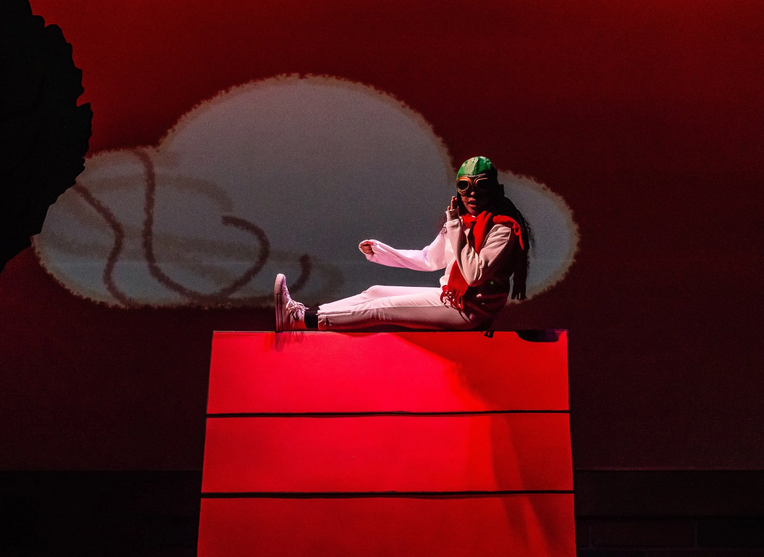 An actor playing Snoopy is sitting on top of a doghouse, wearing goggles, a beige jacket, red scarf, white pants, and sneakers, in front of a background with a large white circular shape and black and red elements.