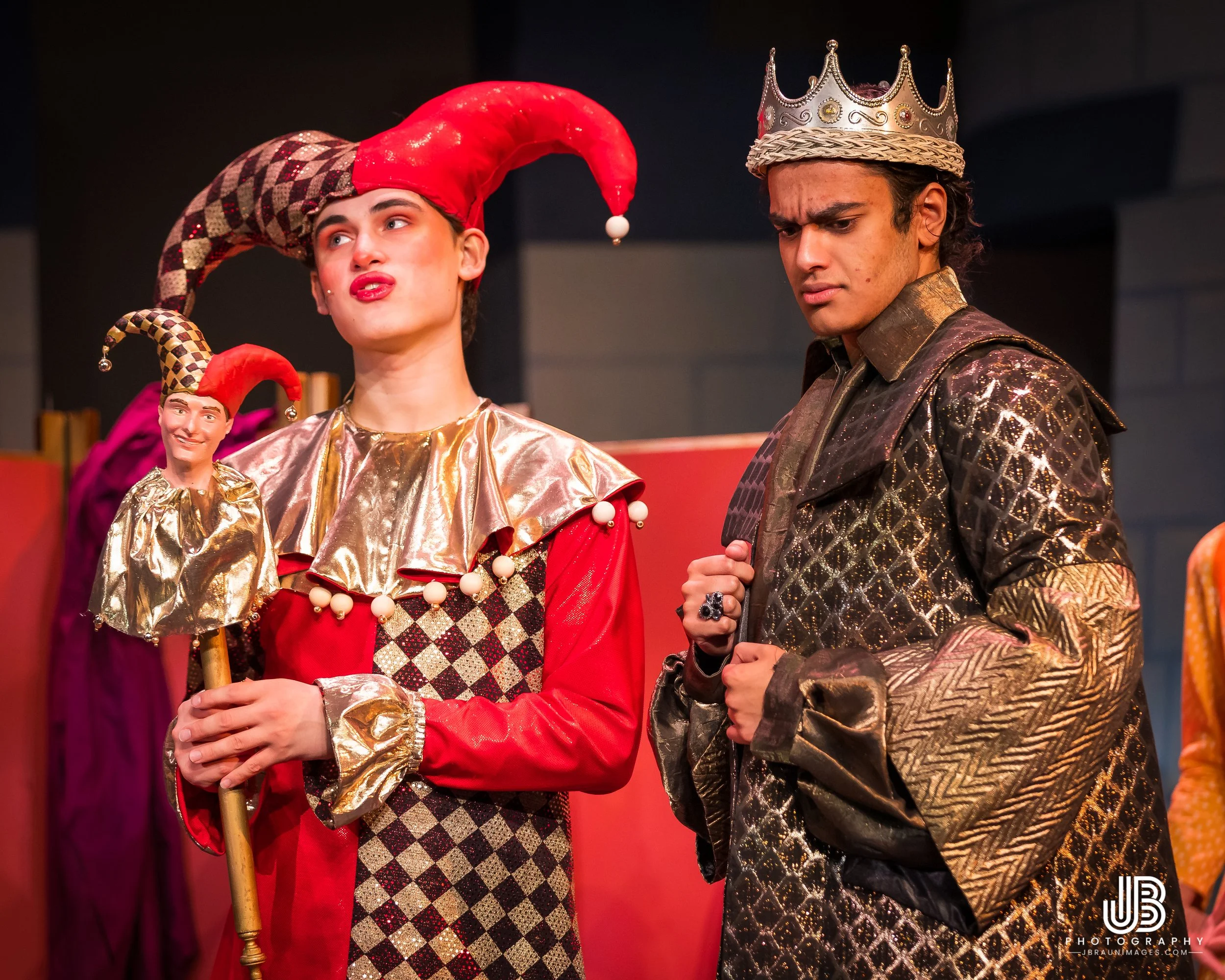 Two people dressed as characters from the play "Once Upon a Mattress", one wearing a red and gold jester outfit with a pointy hat and the other wearing a silver crown and black and gold patterned outfit, standing on stage.