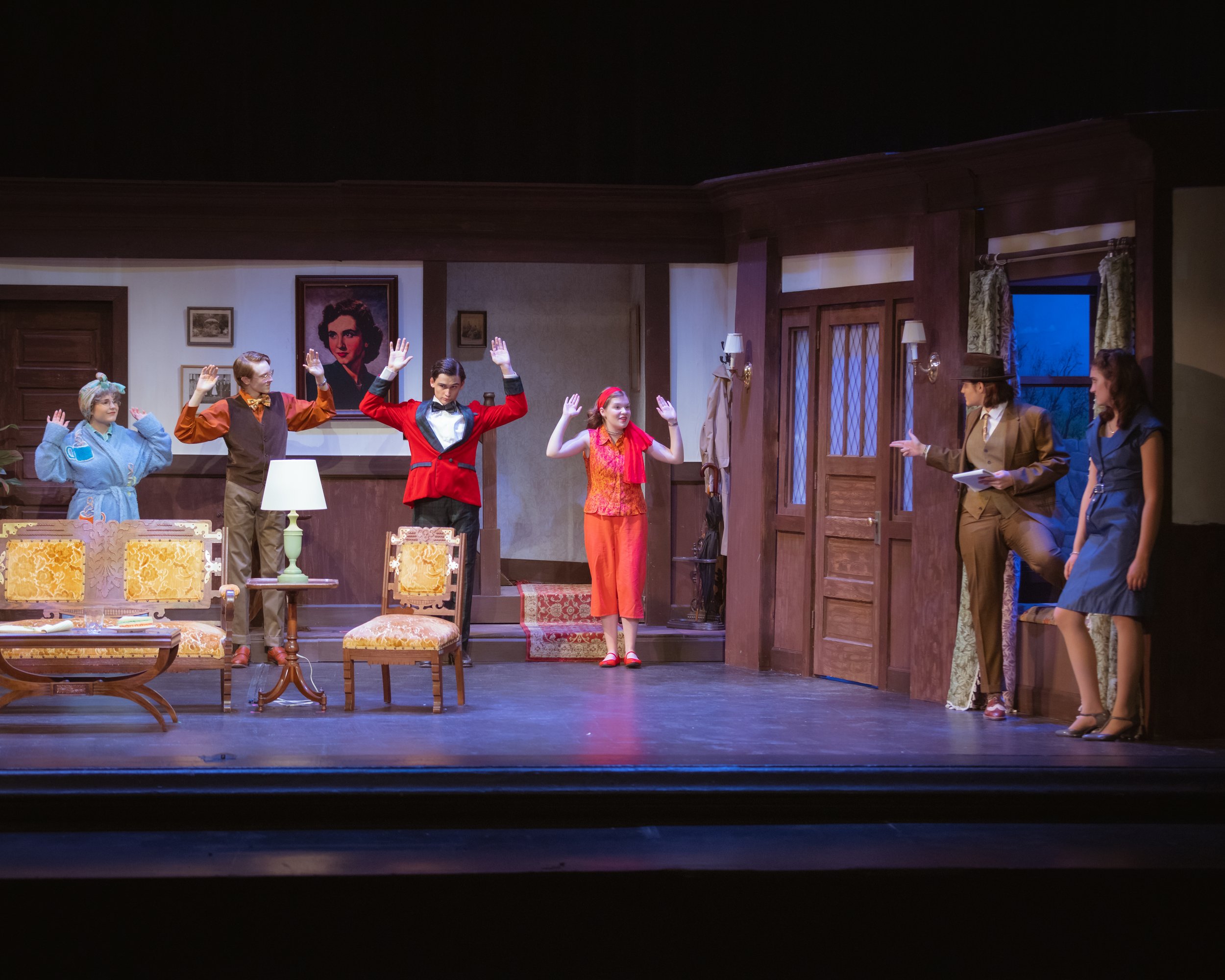The image shows a theatrical stage with seven actors dressed in character costumes, engaged in a performance. The setting includes a cozy interior scene with wooden furniture, artwork on the walls, and a large painting of a woman with curly hair in t