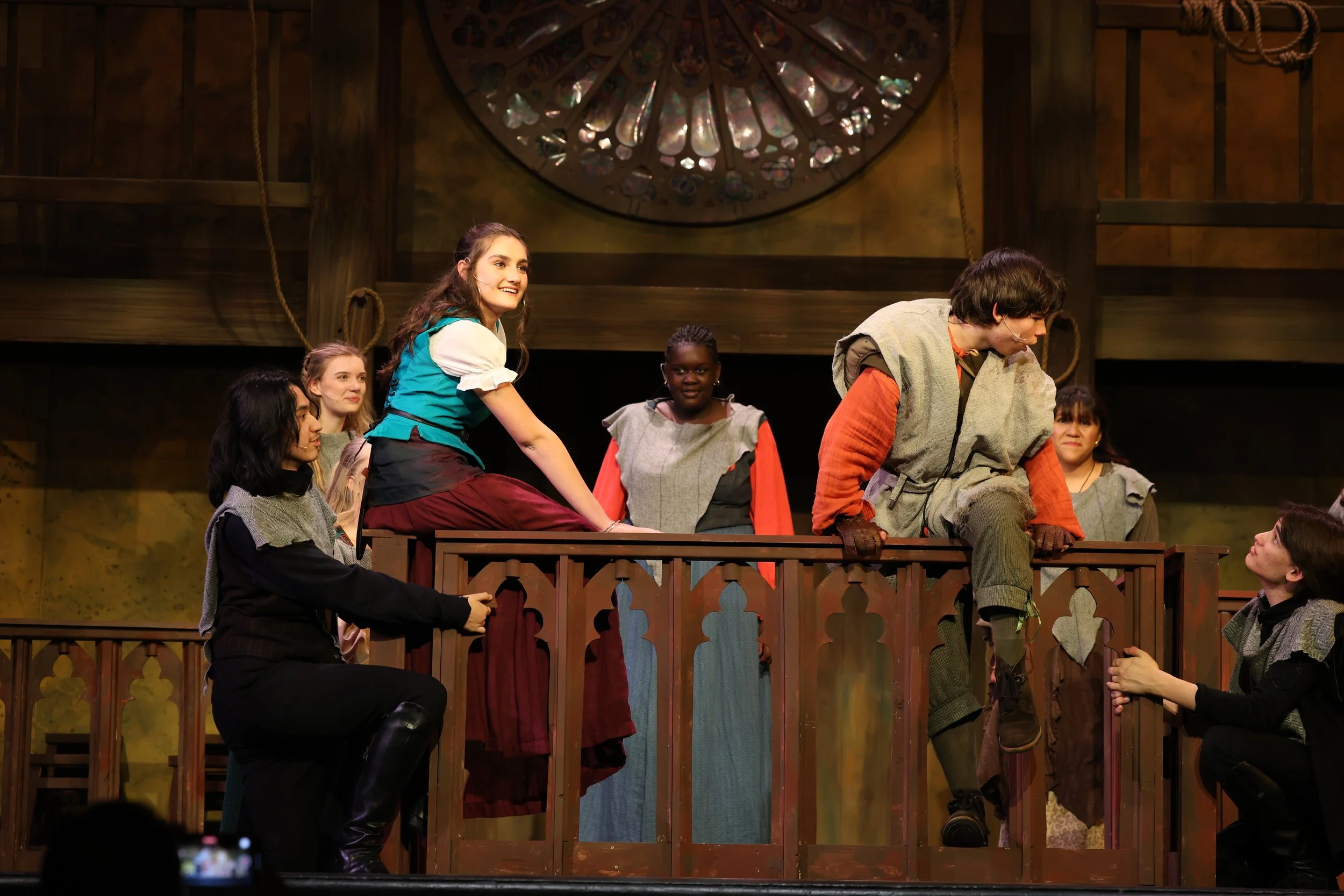 A group of actors in medieval costumes performing on stage with a wooden balcony and stained glass window in the background.
