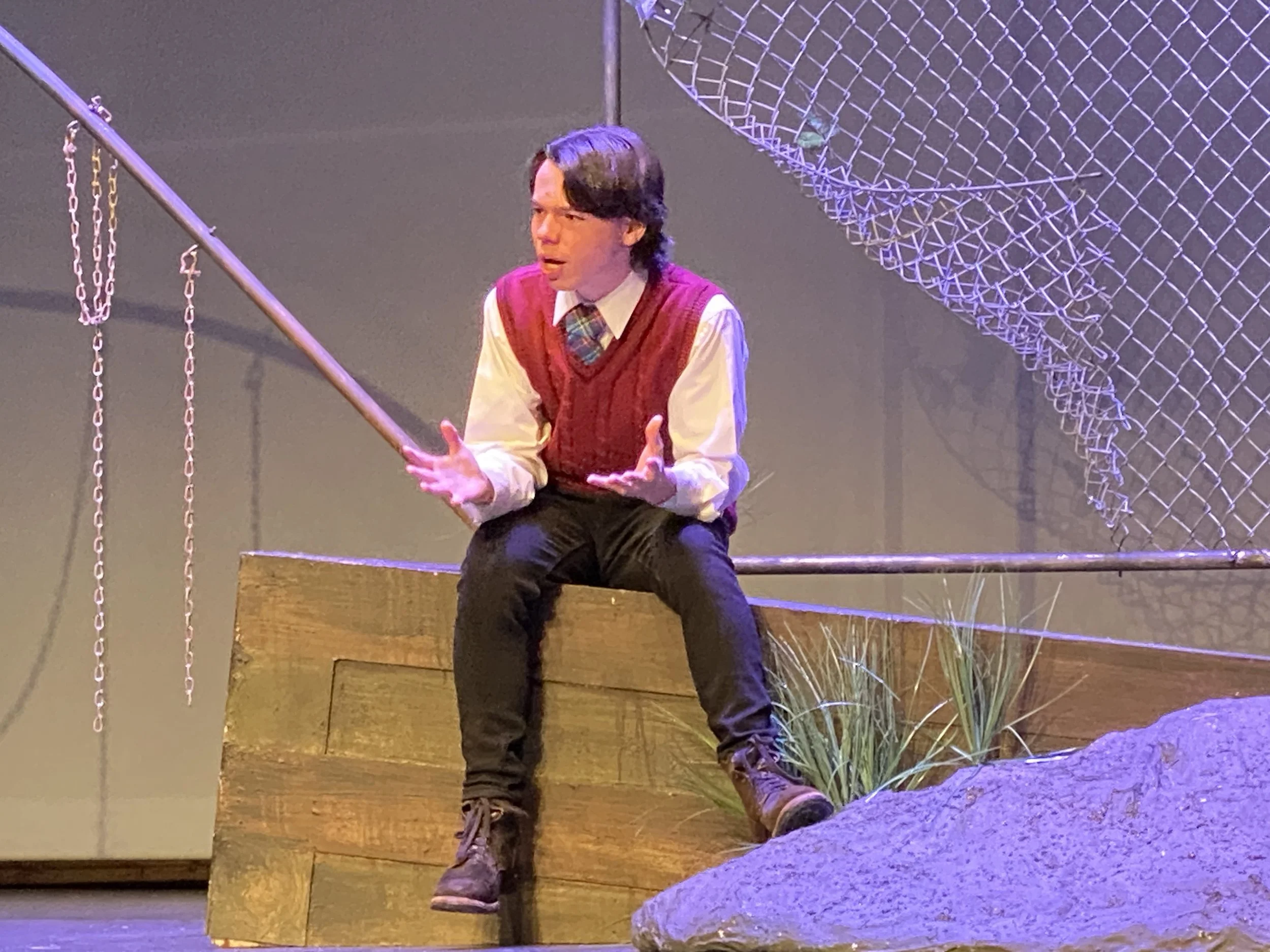 A man with dark hair, wearing a red vest over a white shirt and a plaid tie, is sitting on wooden steps near a chain-link structure, gesturing with his hands during a performance.