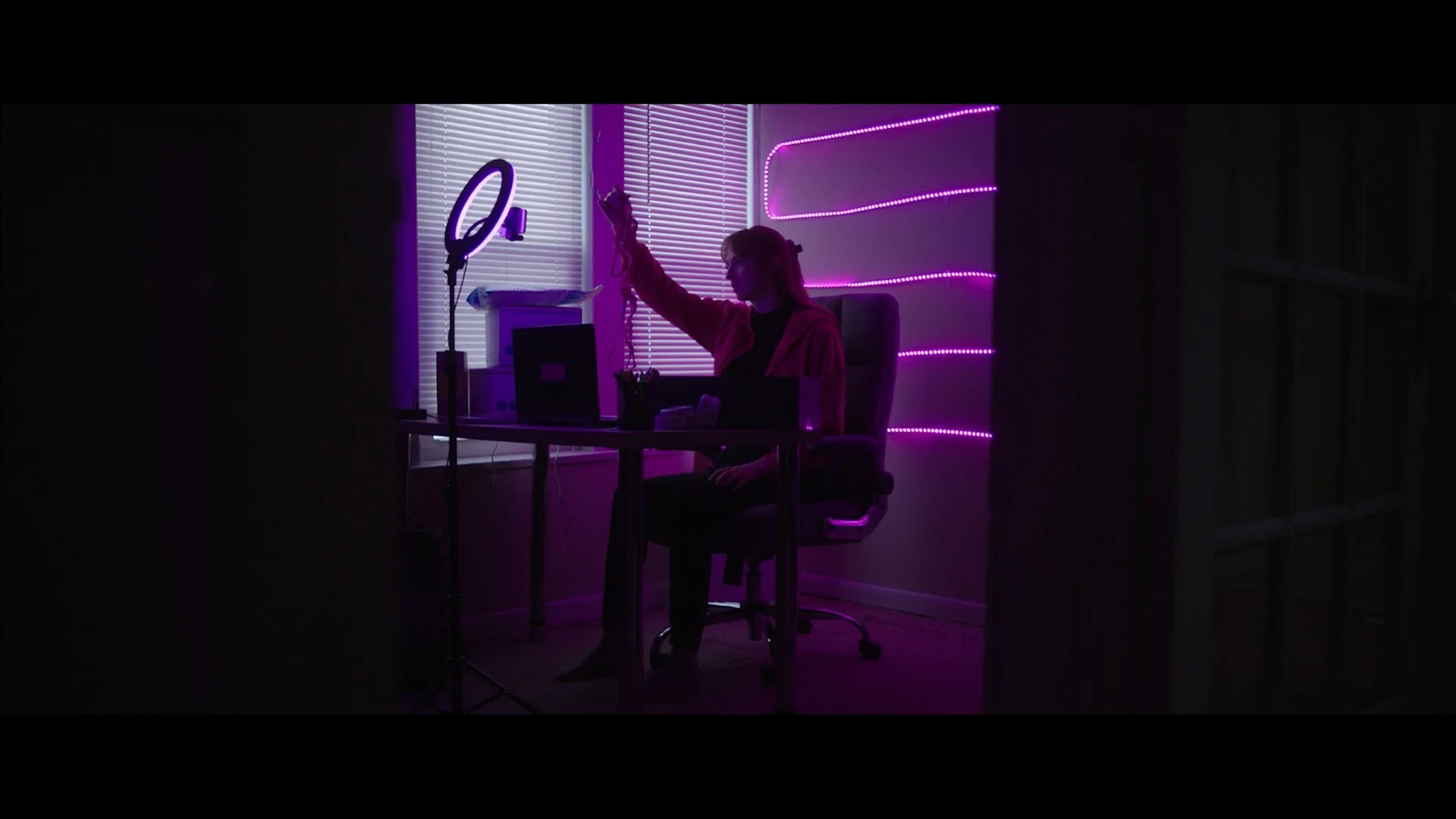 Person sitting at desk in a dark room illuminated by purple LED lights, with blinds on the window and a ring light in front.