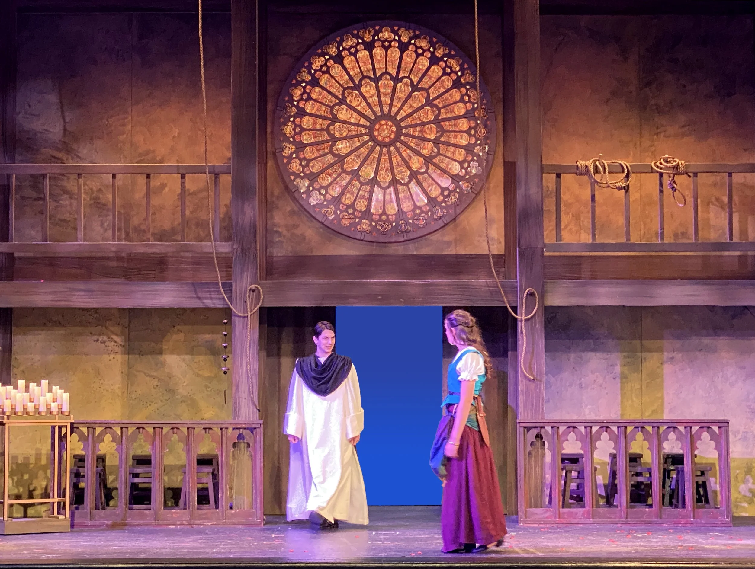 Two actors in historical costumes are performing on a stage with a set resembling a cathedral, featuring wooden balconies, a large circular stained glass window, and candles.