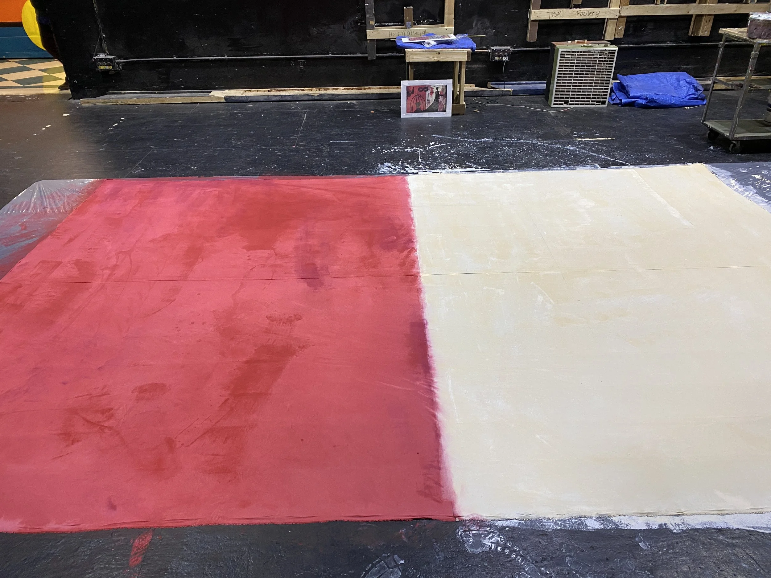 Painted floor with red and beige sections in a workspace or art studio, with various supplies and artwork in the background.