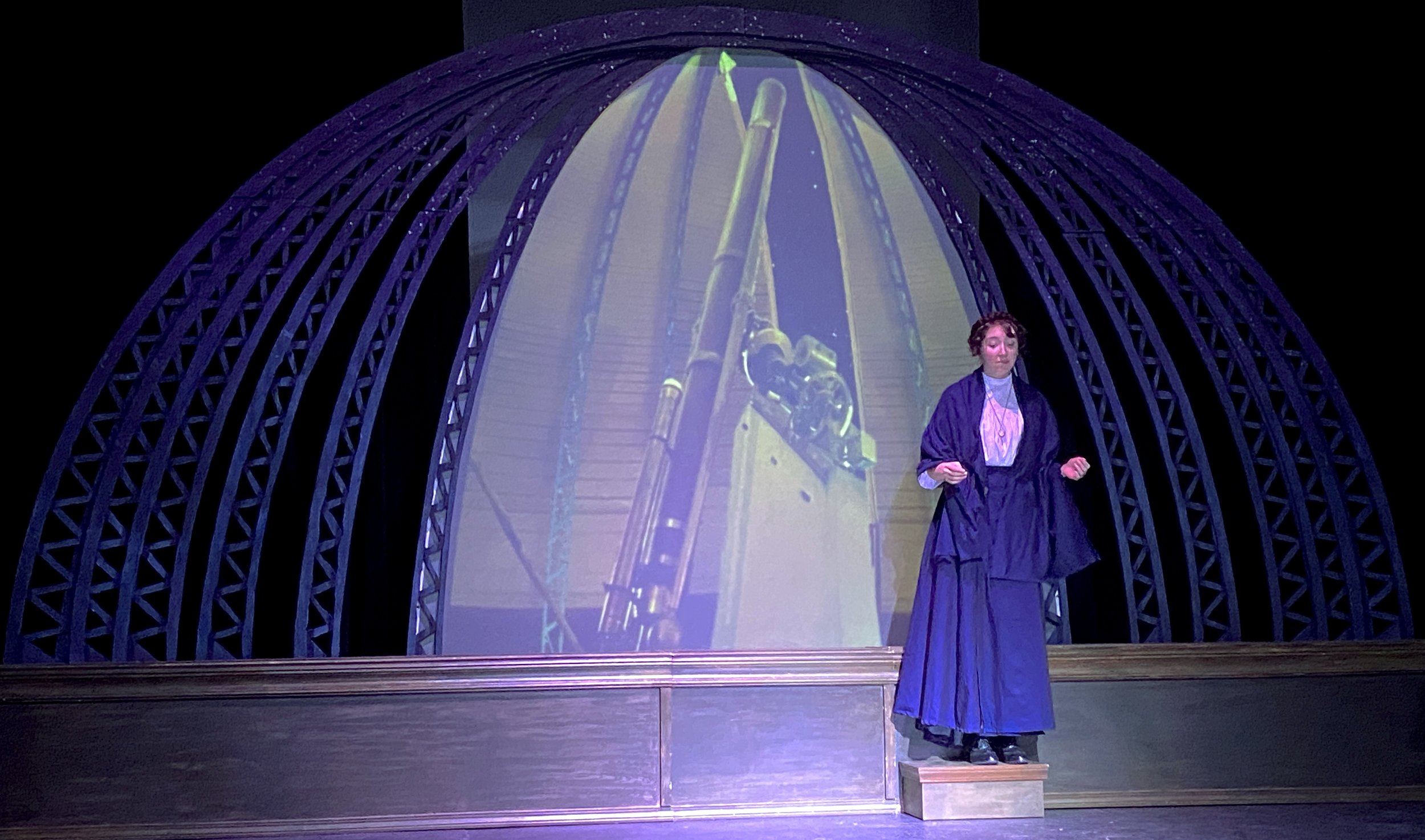 A woman dressed in historical clothing, standing on a small platform, with a large projection of a telescope behind her. The scene is set inside a structure with an arched, lattice-like frame.