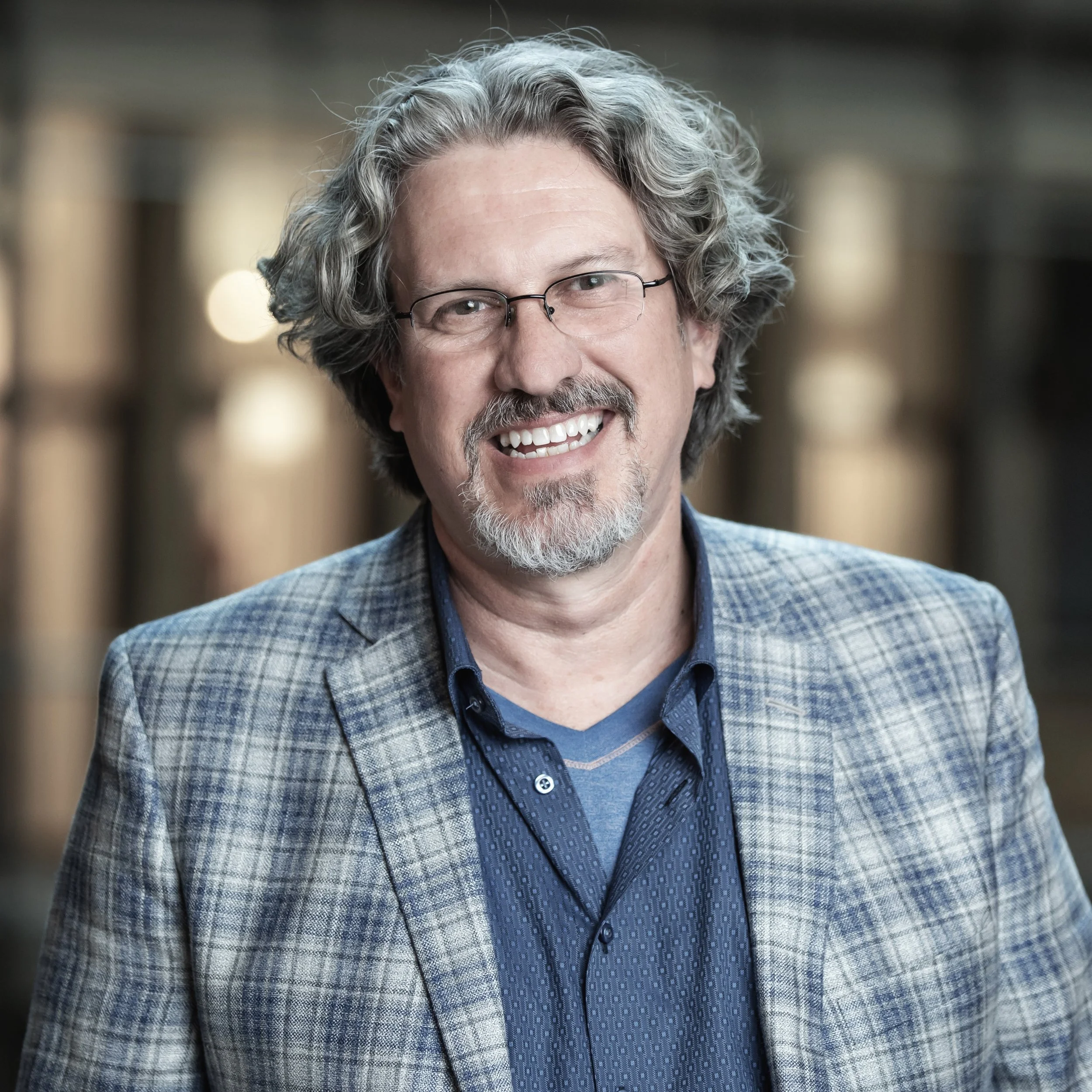 Close-up of a smiling middle-aged man with wavy gray hair, glasses, and a beard, wearing a checked blazer and a blue shirt, in an indoor setting with blurred background lights.
