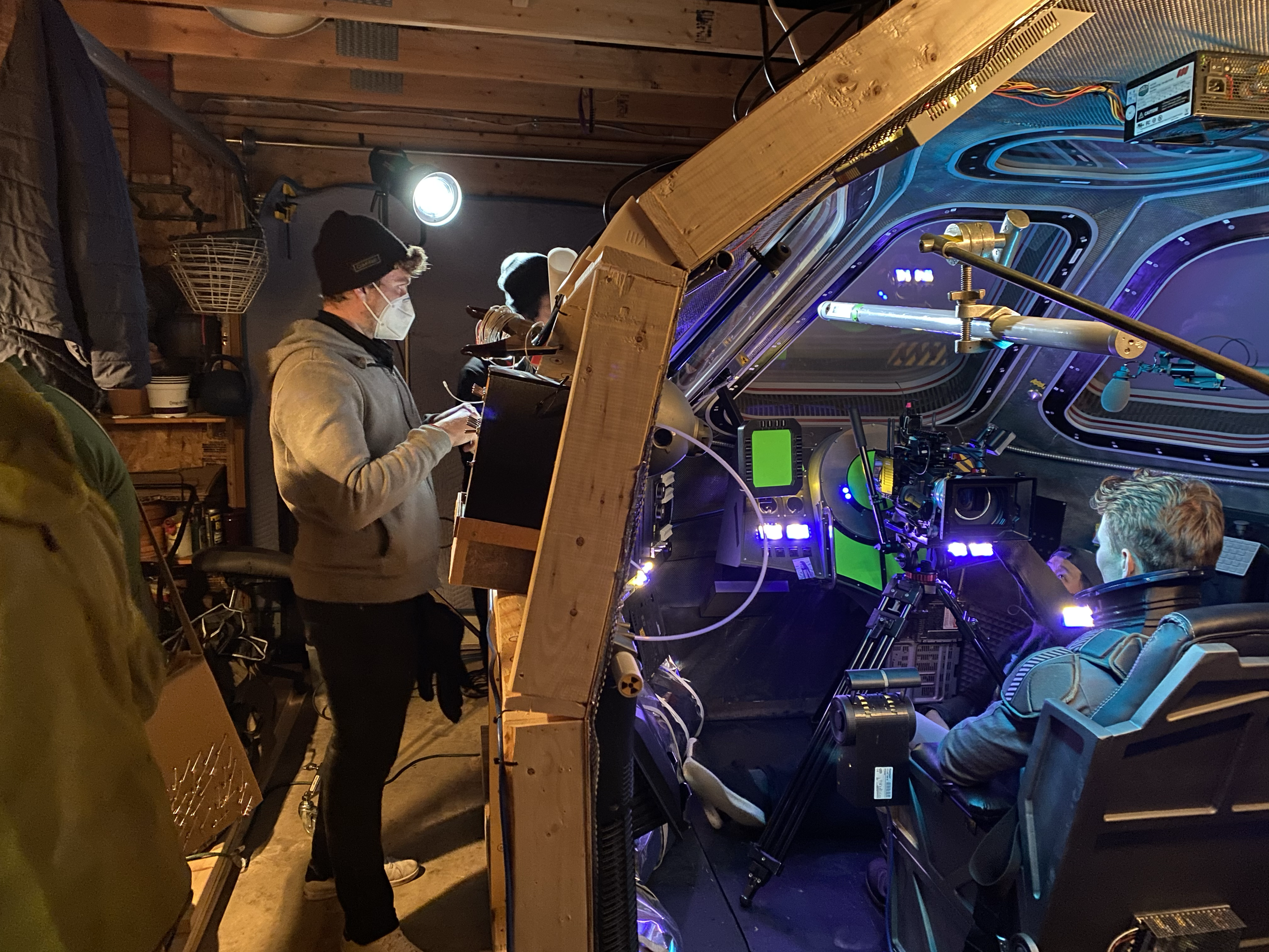 People working on a sound stage set of a spaceship with wooden walls and ceiling. One person is taking notes, others are observing or working on the set. The scenery has colorful, illuminated controls visible.