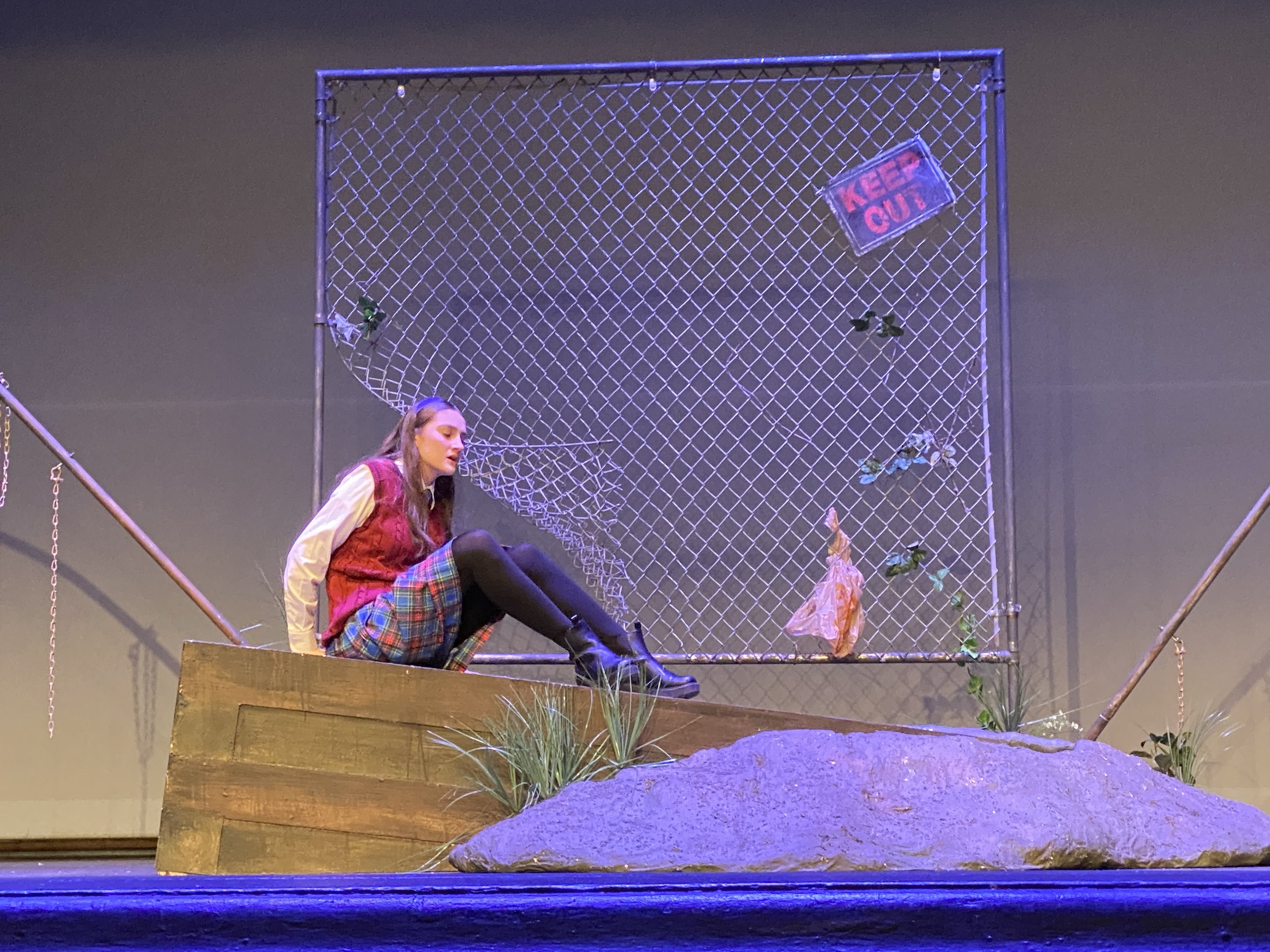 A stage scene with a woman sitting on a wooden platform in front of a chain-link fence backdrop. The fence has a sign that says 'Keep Out' and some artificial plants attached to it. The scene is part of a theatrical performance.