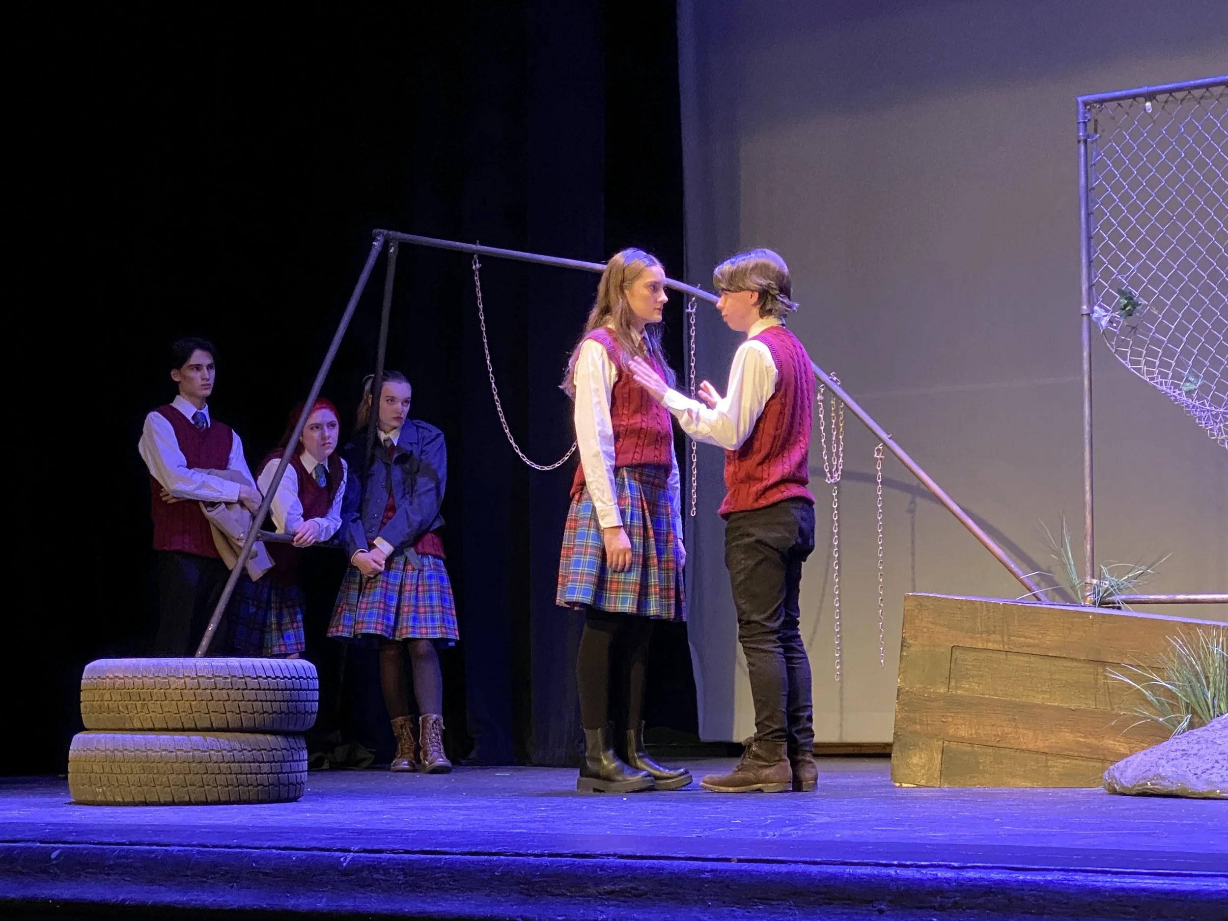 Stage scene from a play with six young actors in school uniforms, two of whom are embracing, while the others watch. Stage props include a tire, a swingset frame, stacked tires, a wooden platform, and a chain link fence.
