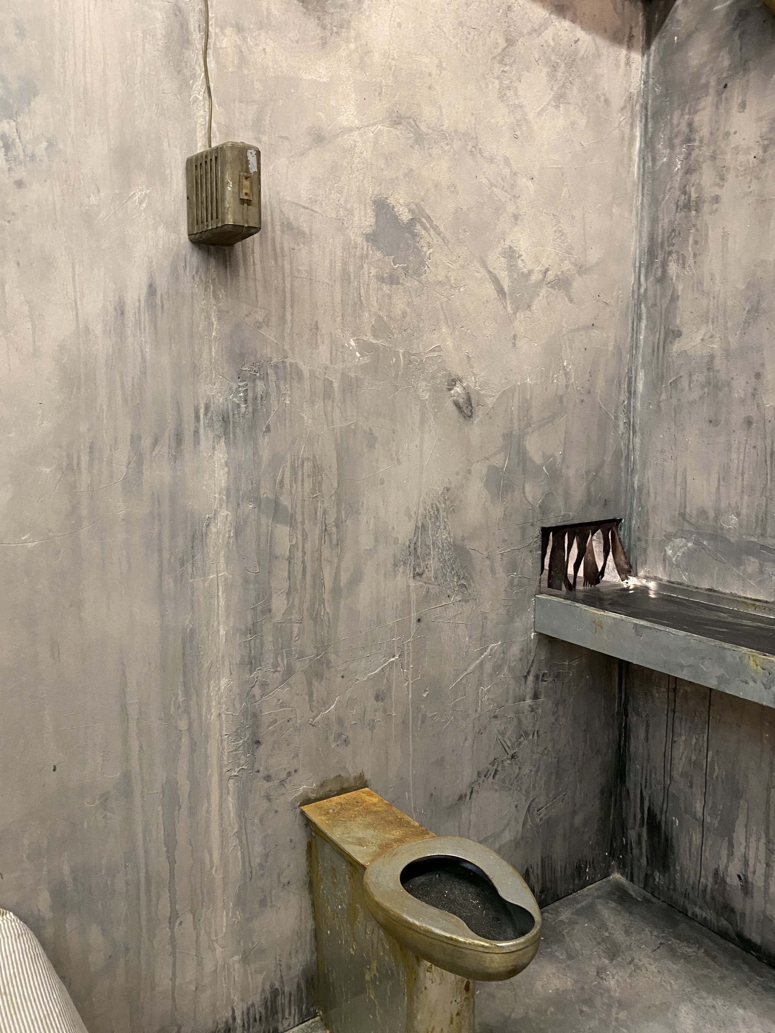 A film set with gray stained walls, a rusty speaker on the wall, a shelf above a modern toilet, and a small grated vent below the shelf.