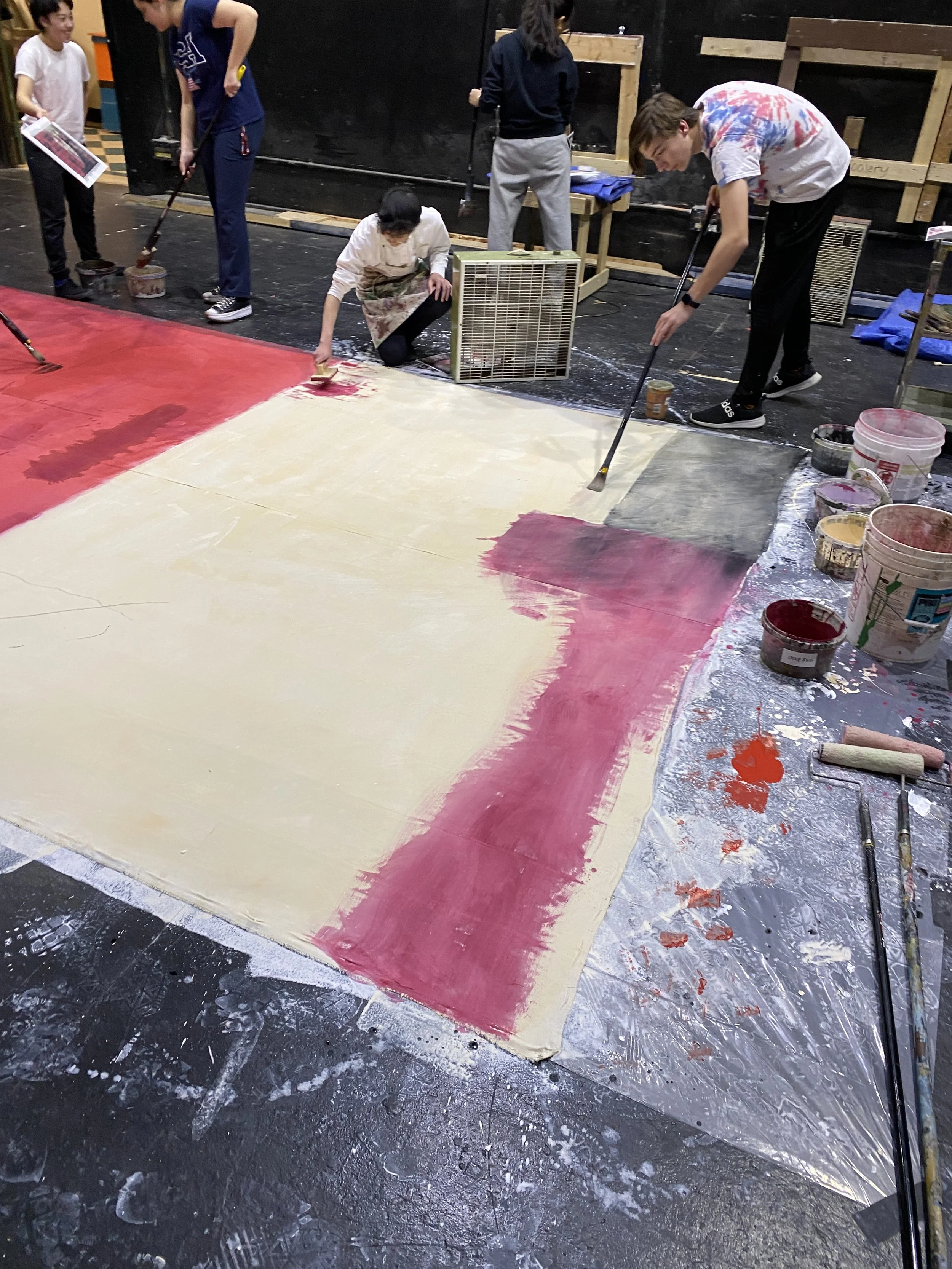 People painting a large canvas with red and white paint, surrounded by paint buckets and brushes, in an indoor setting.