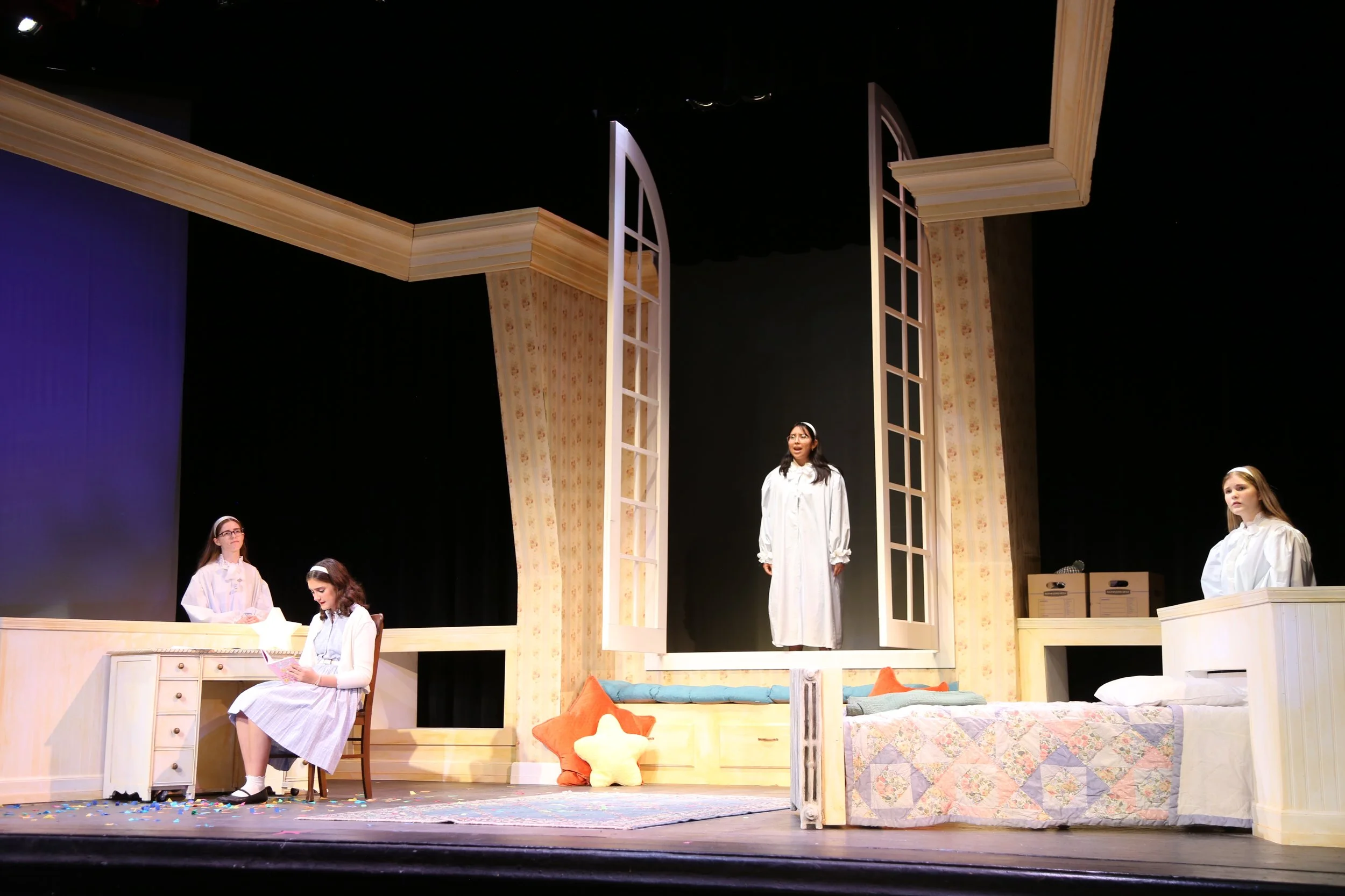 A scene from a stage play featuring five young women dressed in vintage nightgown costumes, performing on a set designed to look like a cozy bedroom with beds, furniture, and decorative accents.