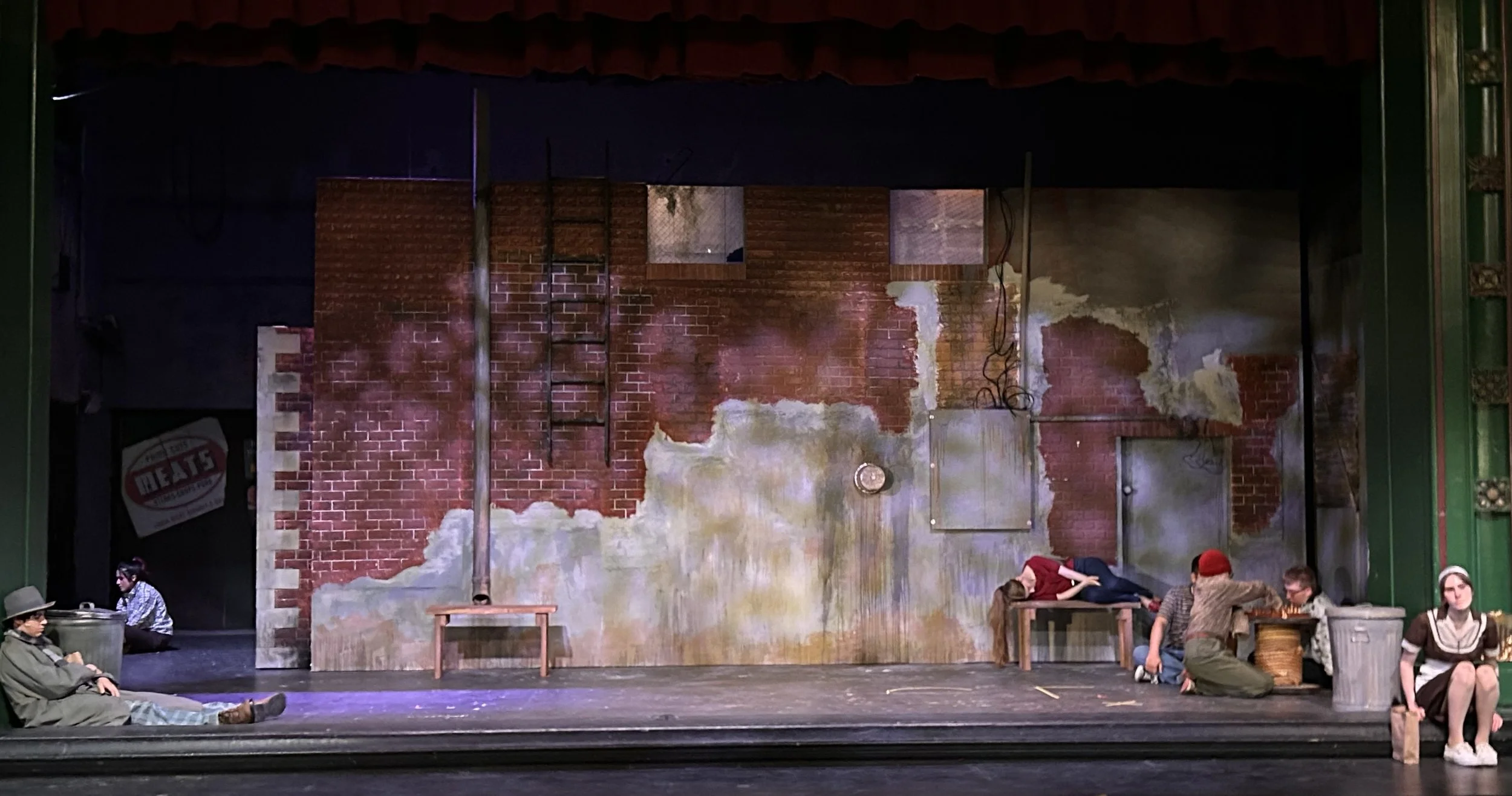 The image shows a theatrical stage set resembling a weathered brick wall, with actors sitting and lying on the stage floor in front of it. Some actors are sitting near a trash can and a picnic basket, while others are lying down.