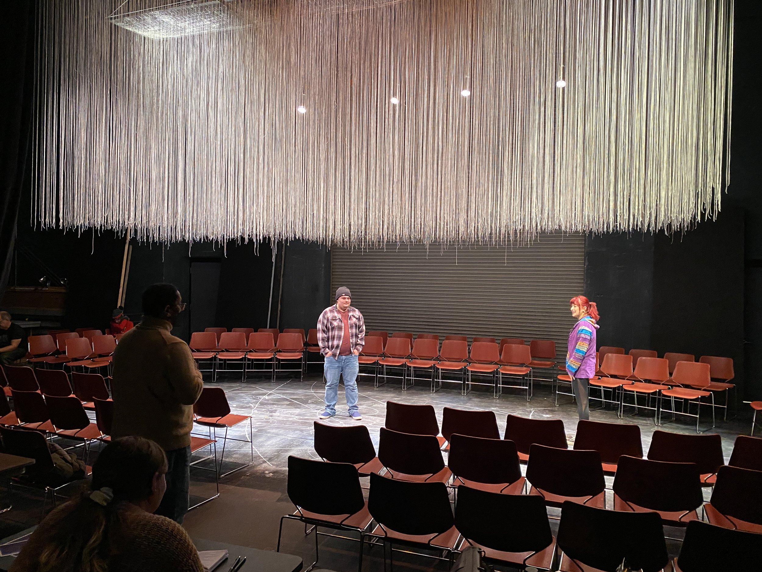 People rehearsing a play in an auditorium with a large hanging installation of strings on the ceiling.