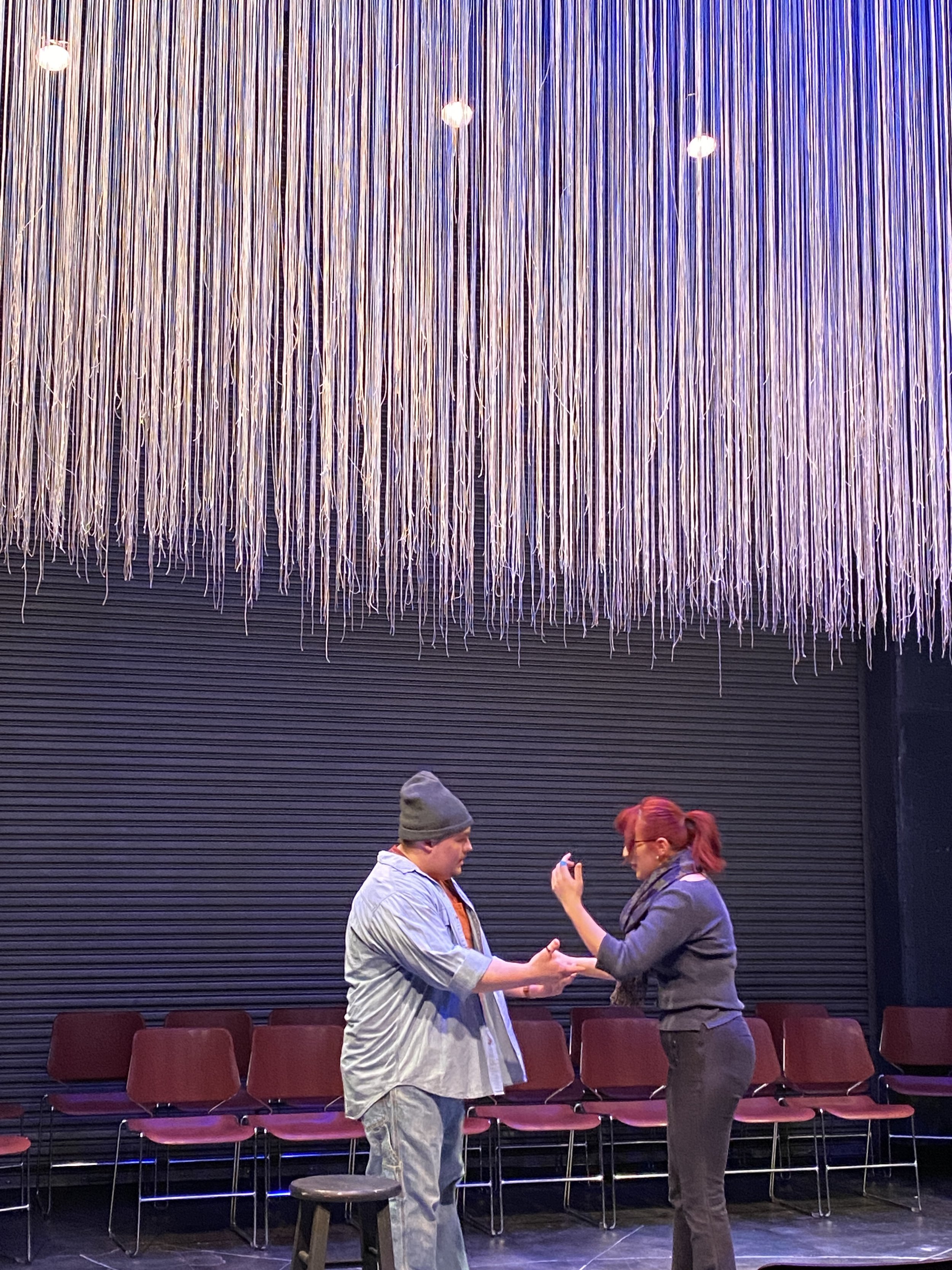 A man and woman are standing and shaking hands in front of a row of empty chairs. They are engaging in a conversation, with the woman holding a phone. The background features a decorative hanging string installation.