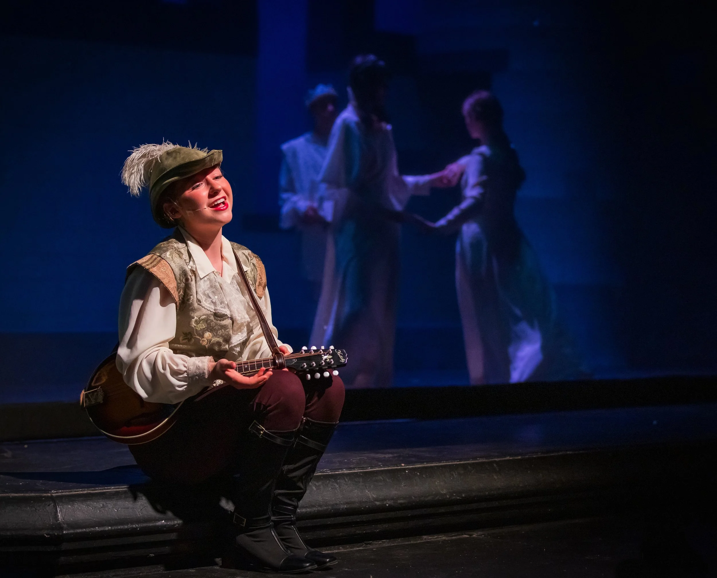 A woman dressed in historical costume with a feathered hat, smiling and playing a guitar on stage, with a dark background and dim lighting.