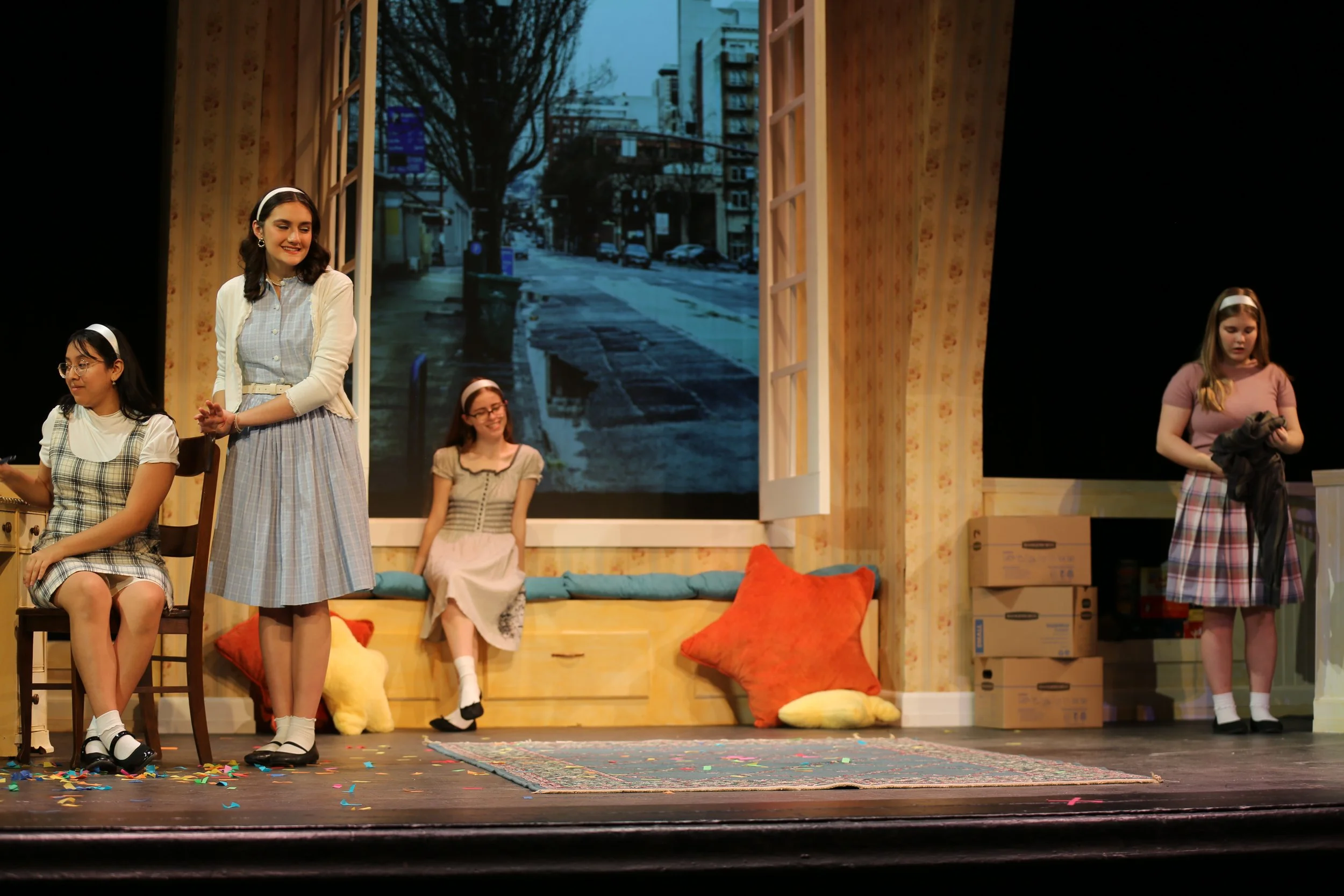 A stage scene with five young women in vintage-style clothing, some sitting and some standing, with a backdrop of a city street scene and colorful pillows on the floor.
