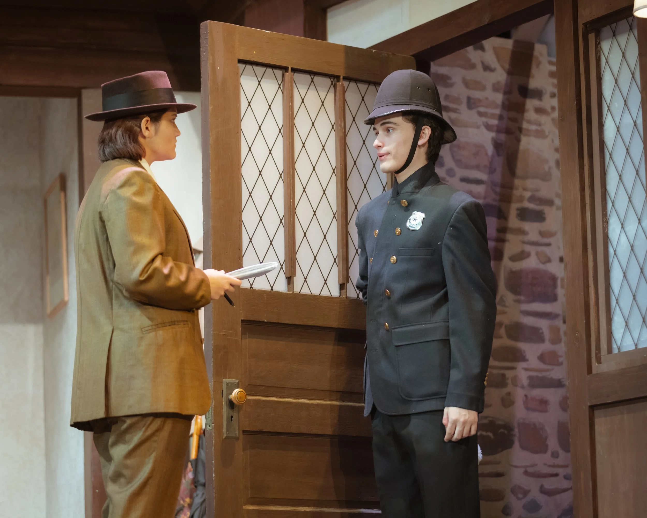 A stage set with a detective in a beige blazer and wide-brimmed hat talking to a police officer in a dark uniform and helmet inside a wooden structure with a brick wall background.