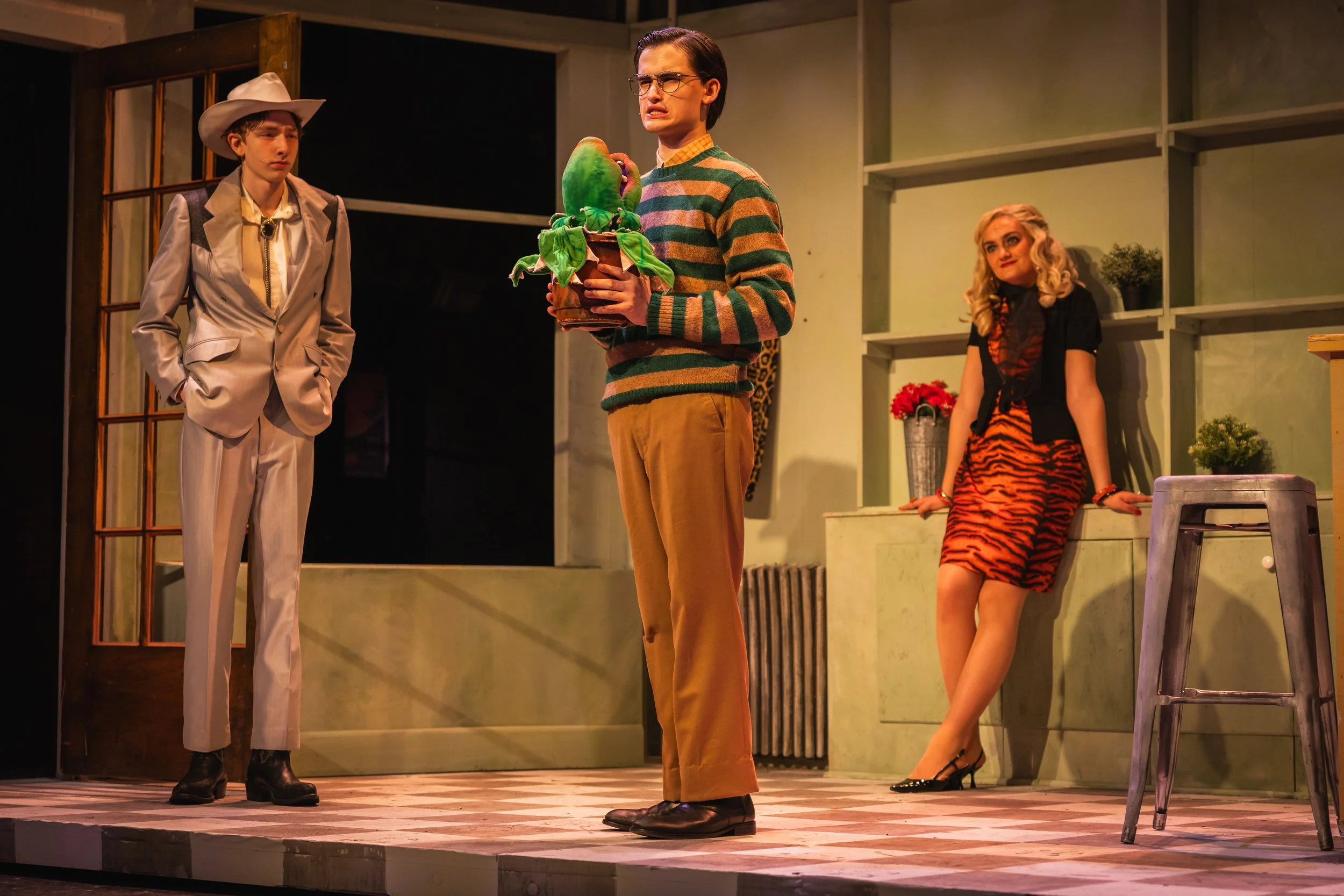 Three actors on stage in a play, with a woman in tiger-print dress sitting on a bench, a man in a striped sweater holding a plant, and a man in a light-colored suit and cowboy hat looking serious.